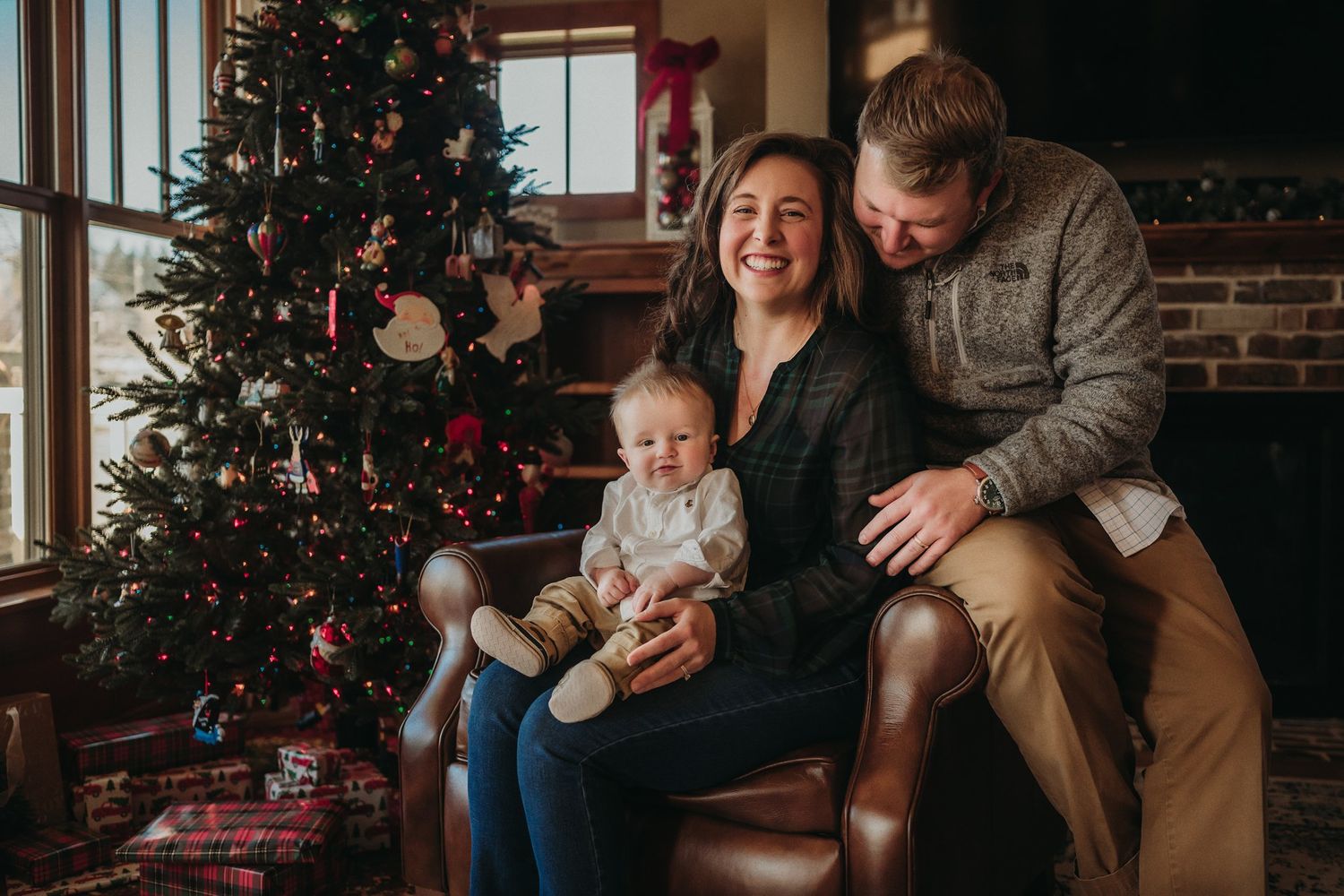 Family poses together by decorated Christmas tree near brick fireplace.