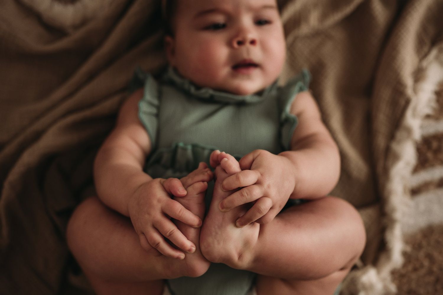 Close-up portrait of a baby playing with their toes on a beige blanket.