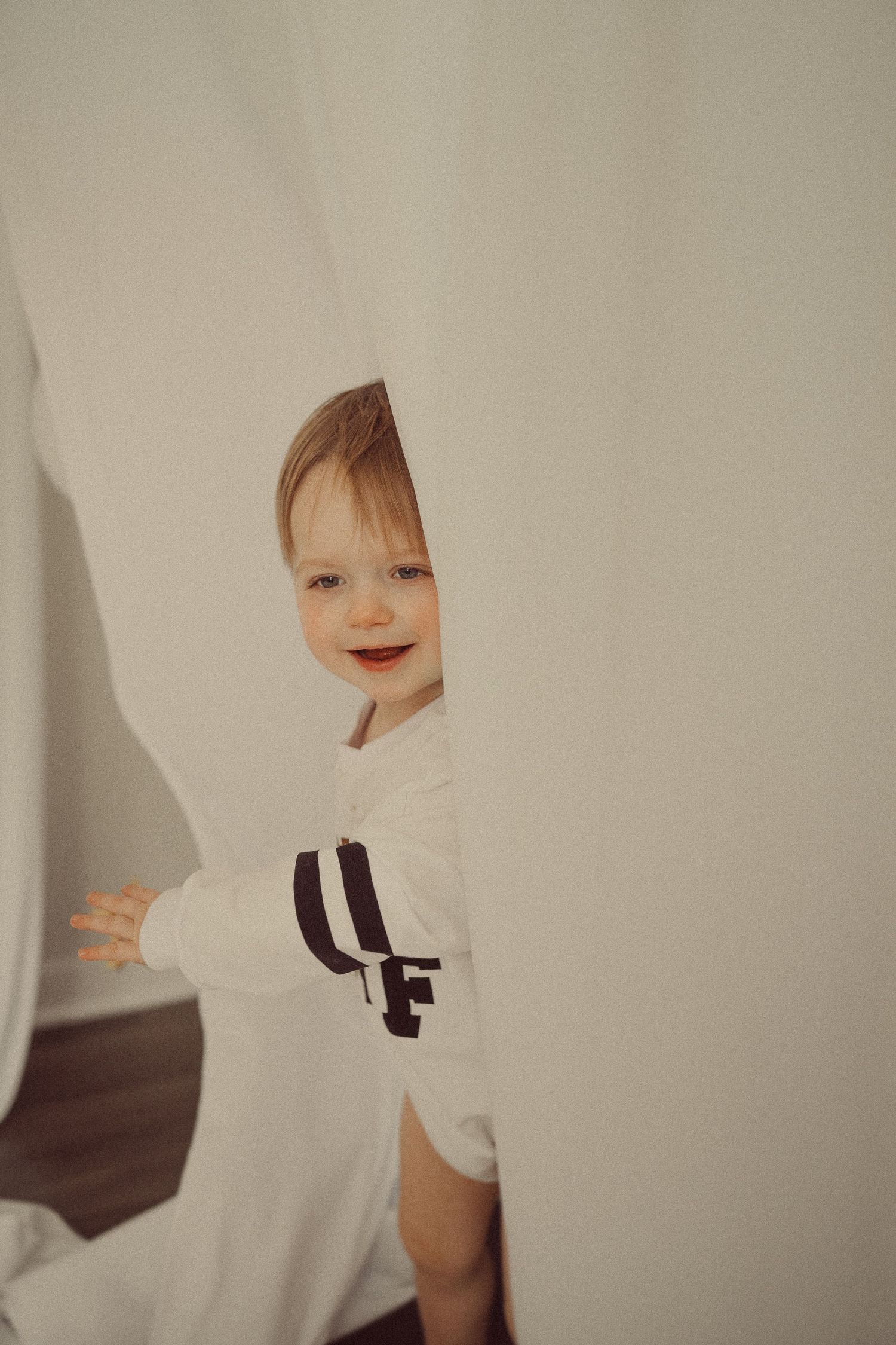 Young child peeking playfully from behind white fabric while wearing a sporty shirt.