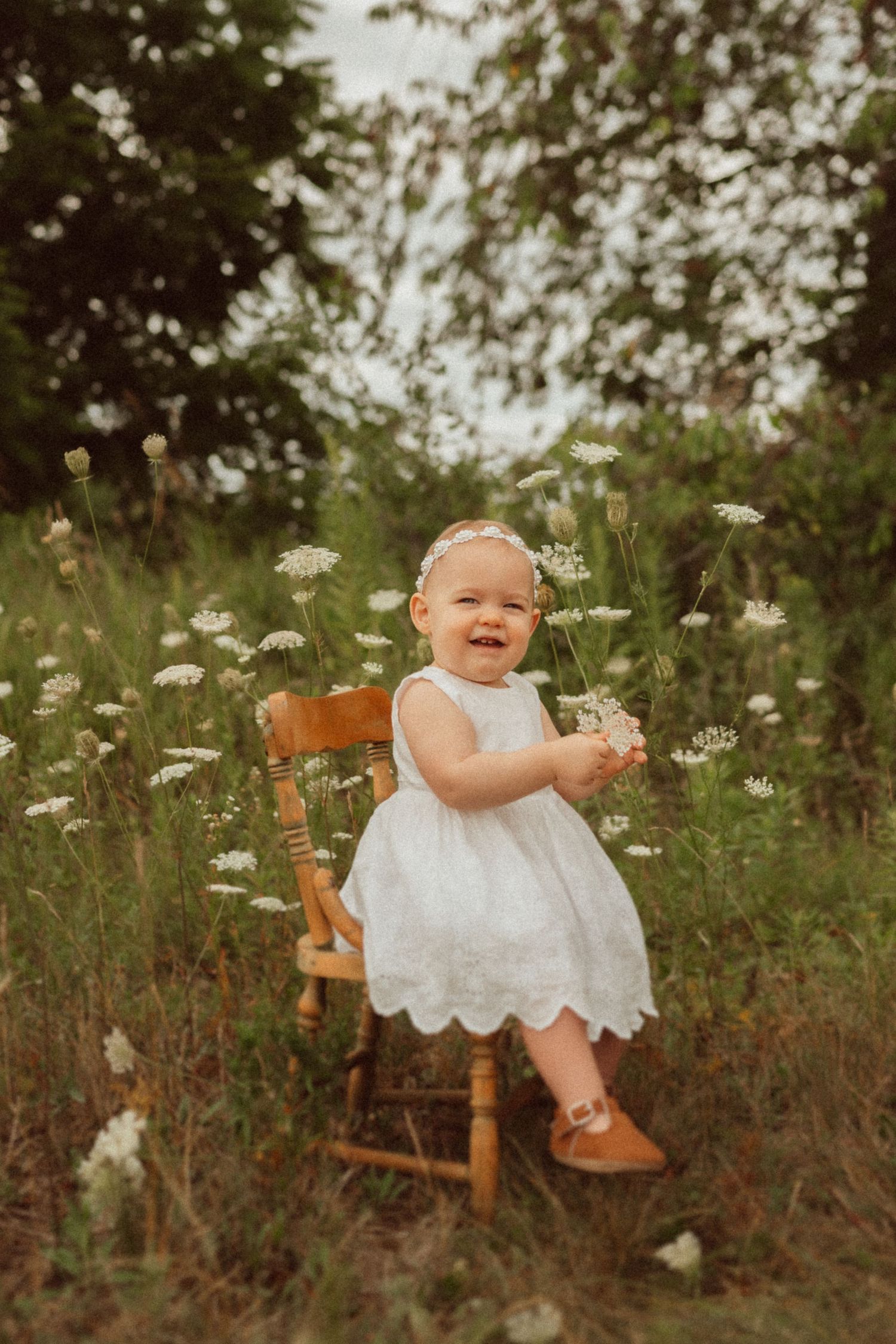 Baby in white dress sits on wooden chair surrounded by blooming wildflowers.