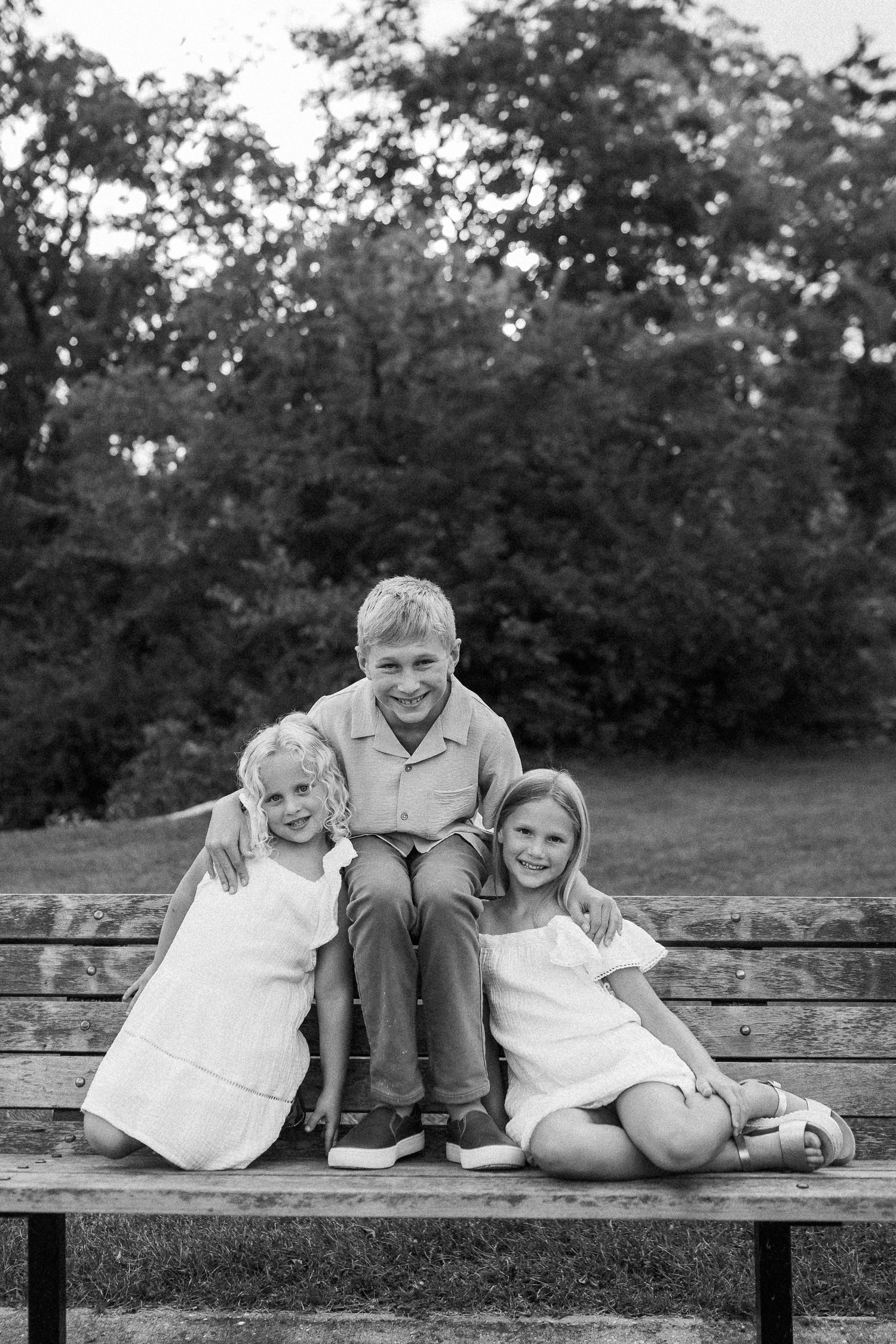 Three children sit together on a wooden bench in a black and white outdoor portrait.