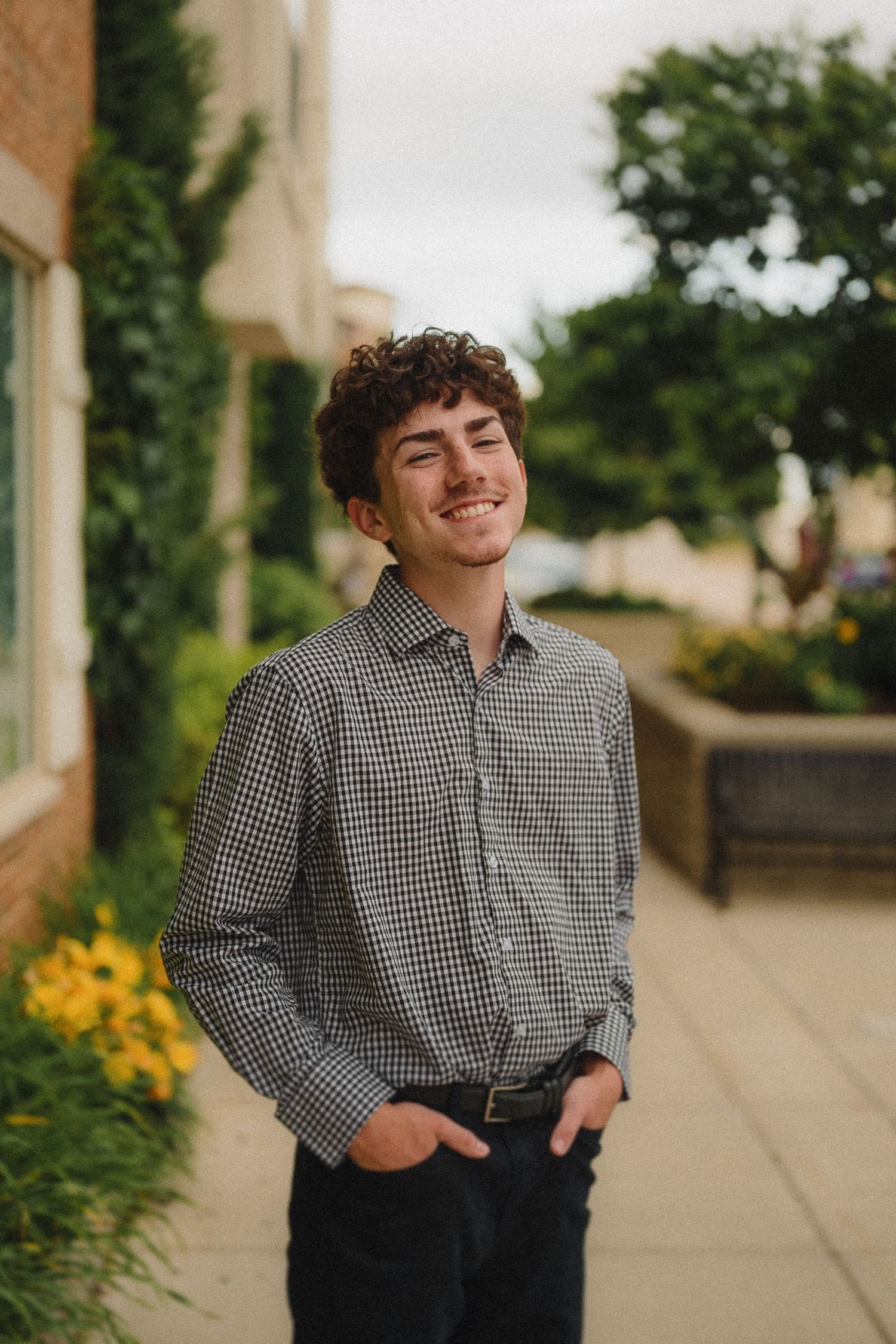 A person in a checkered button down shirt and dark pants stands on a sidewalk with yellow flowers in the background.