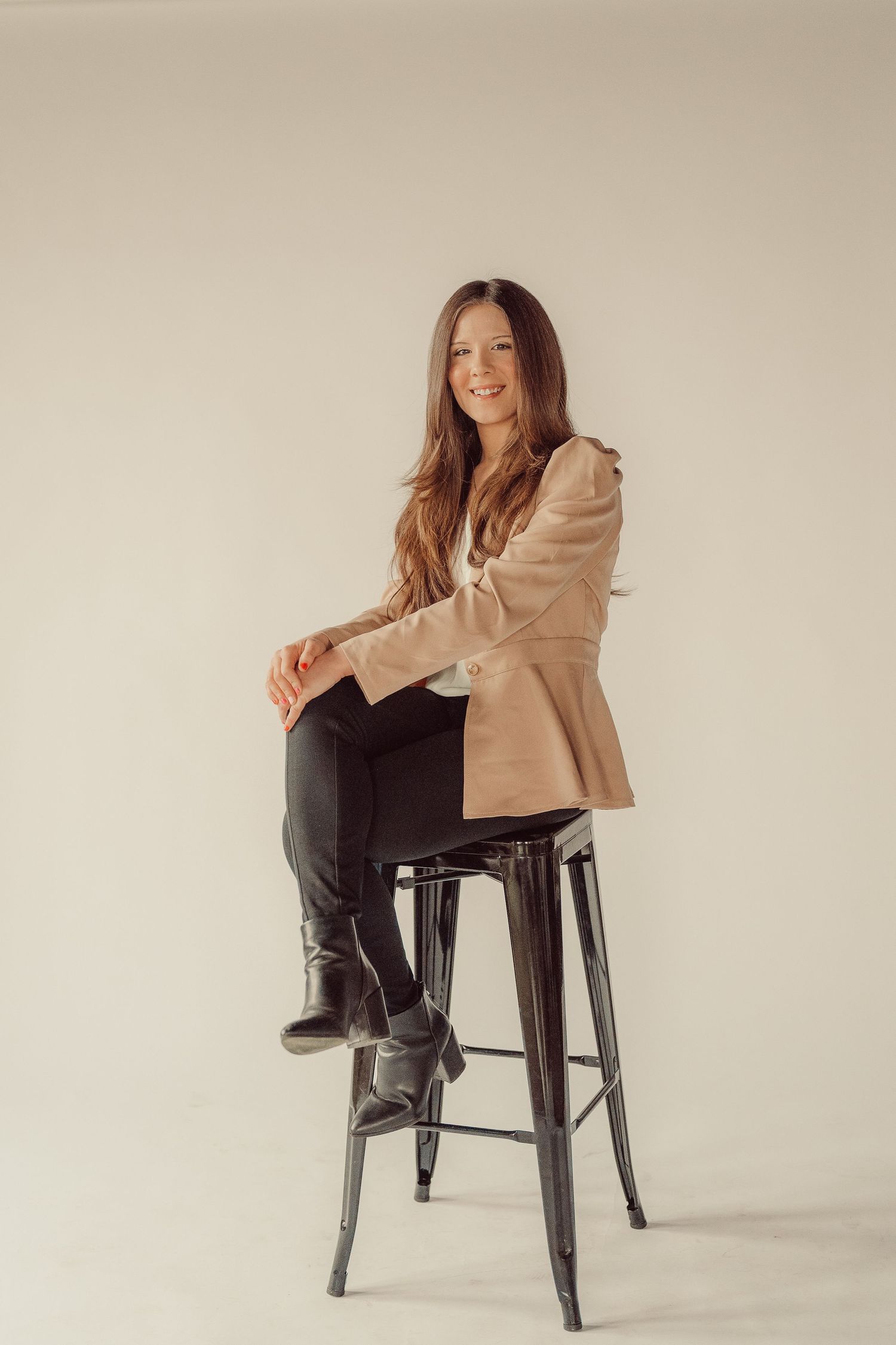 Fashion portrait of woman in beige blazer and black boots sitting on metal stool.