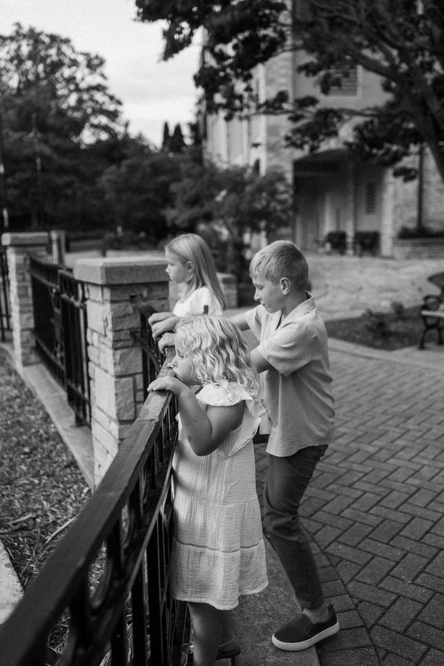 Playful candid moments of children exploring and leaning on a stone wall with iron railing on a brick pathway.