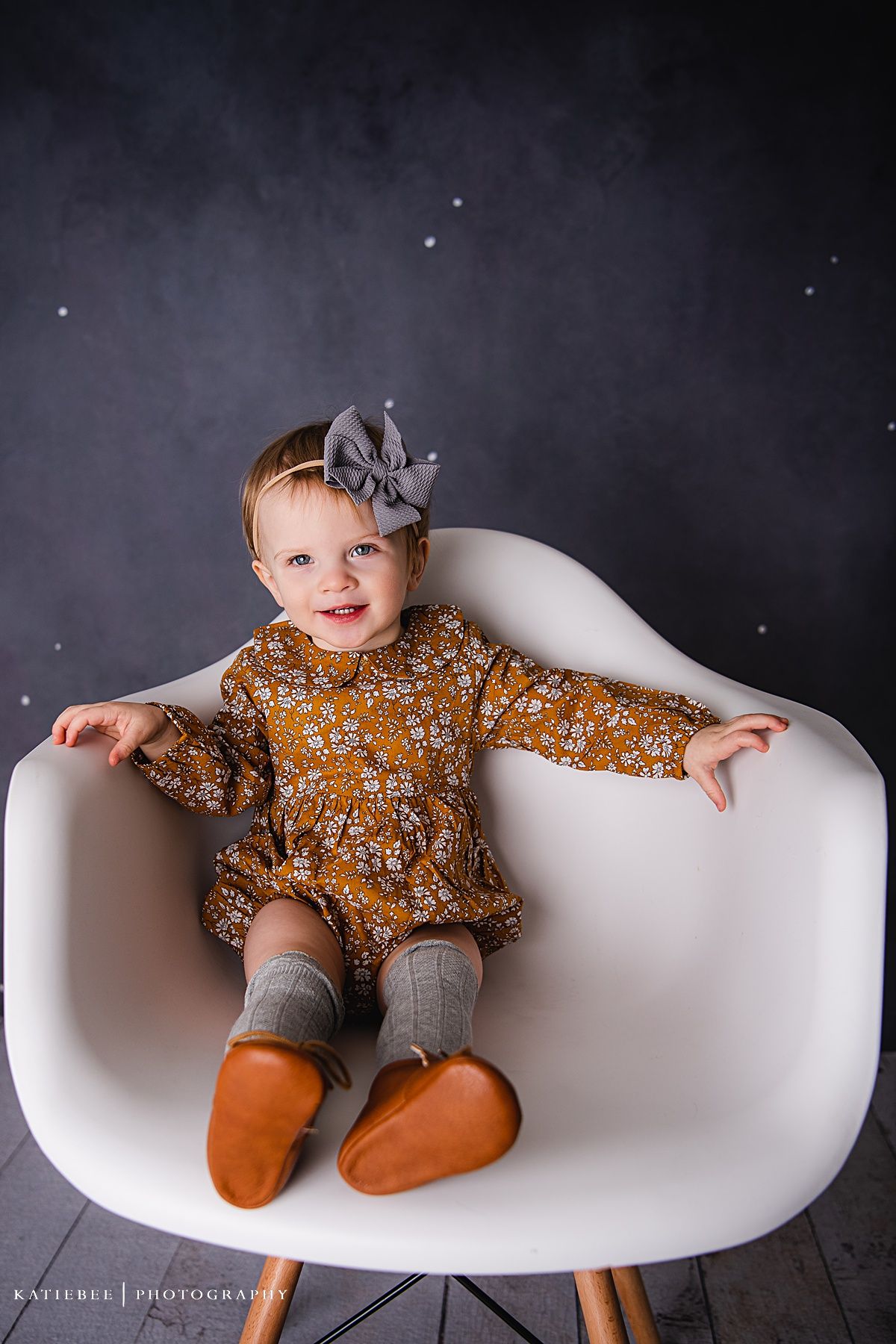 A child in a mustard floral dress and grey knee socks sits comfortably in a white designer chair.