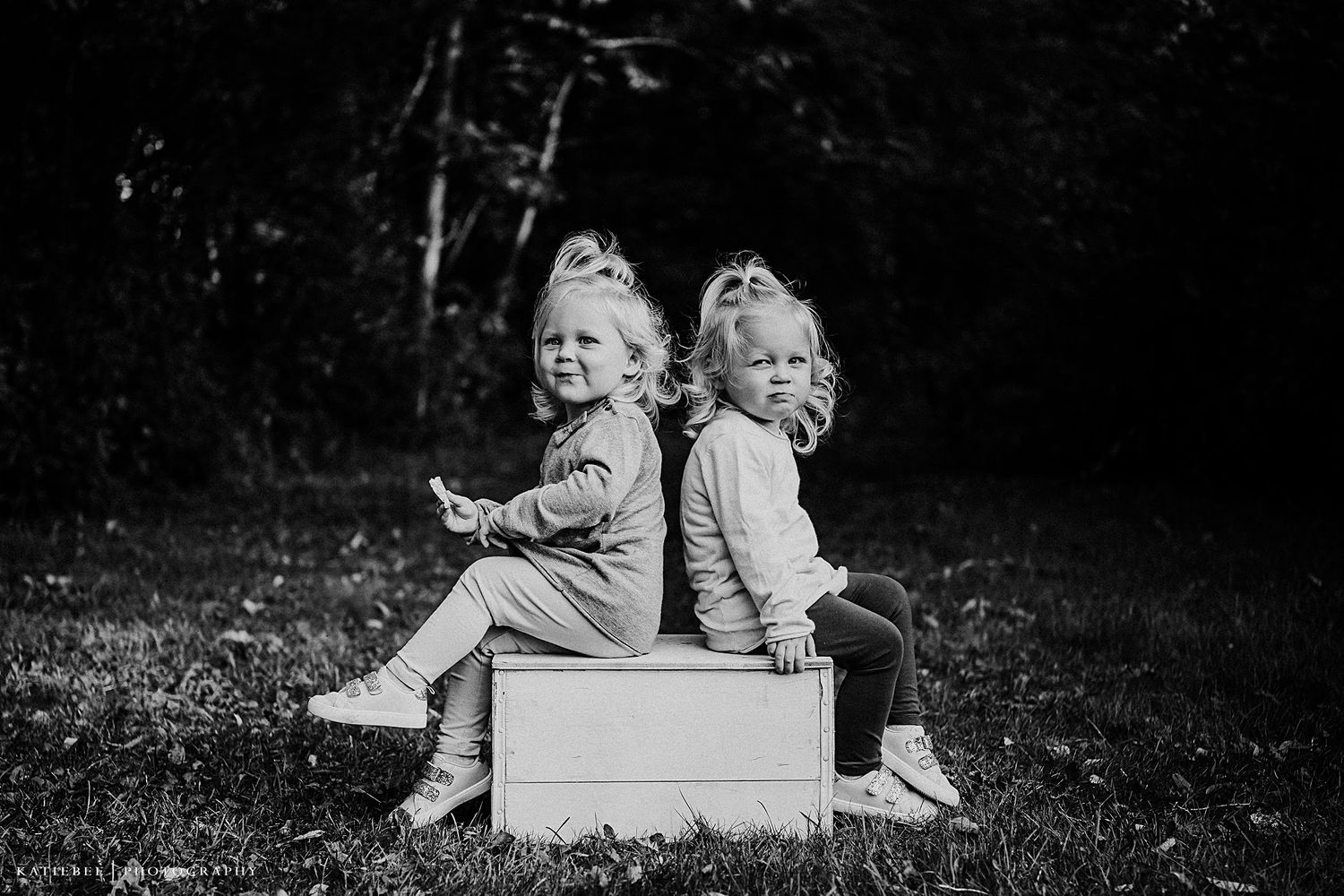 Two children sit back-to-back on a white box, sharing laughter in black and white.
