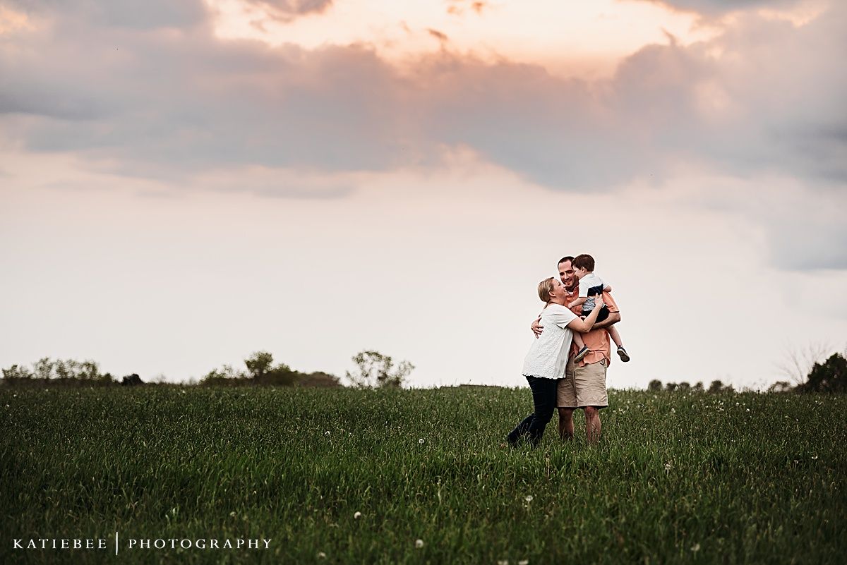 A romantic sunset portrait of a couple embracing in a green meadow against a pink sky.