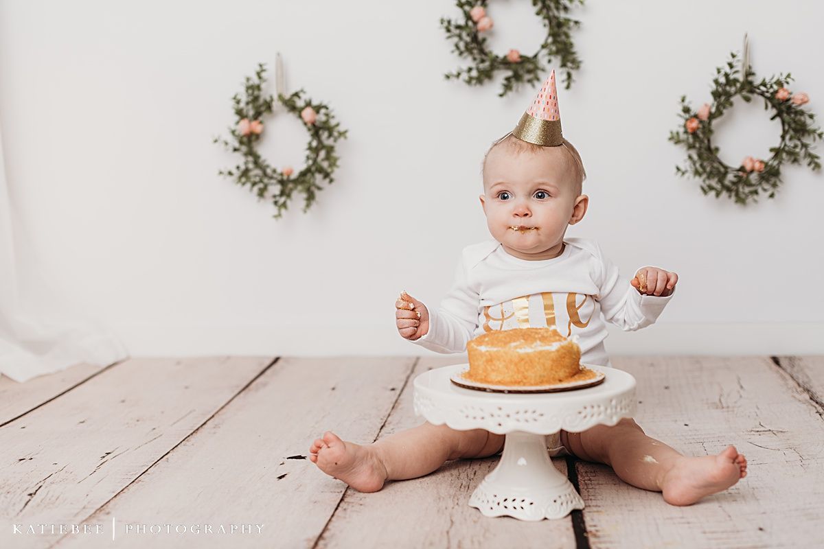 Smiling baby in white onesie next to cake with decorative wreaths hanging on wall.