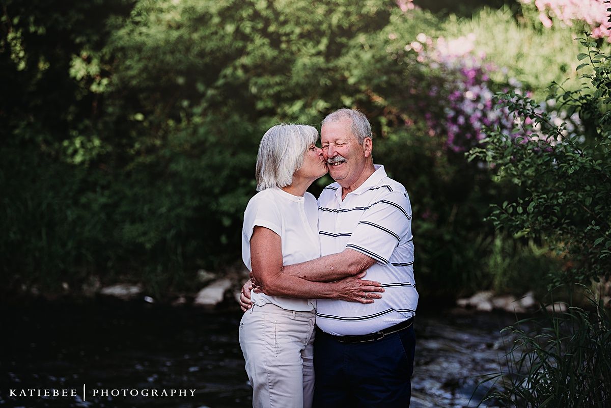 Mature couple embracing outdoors near flowering bushes and stream.