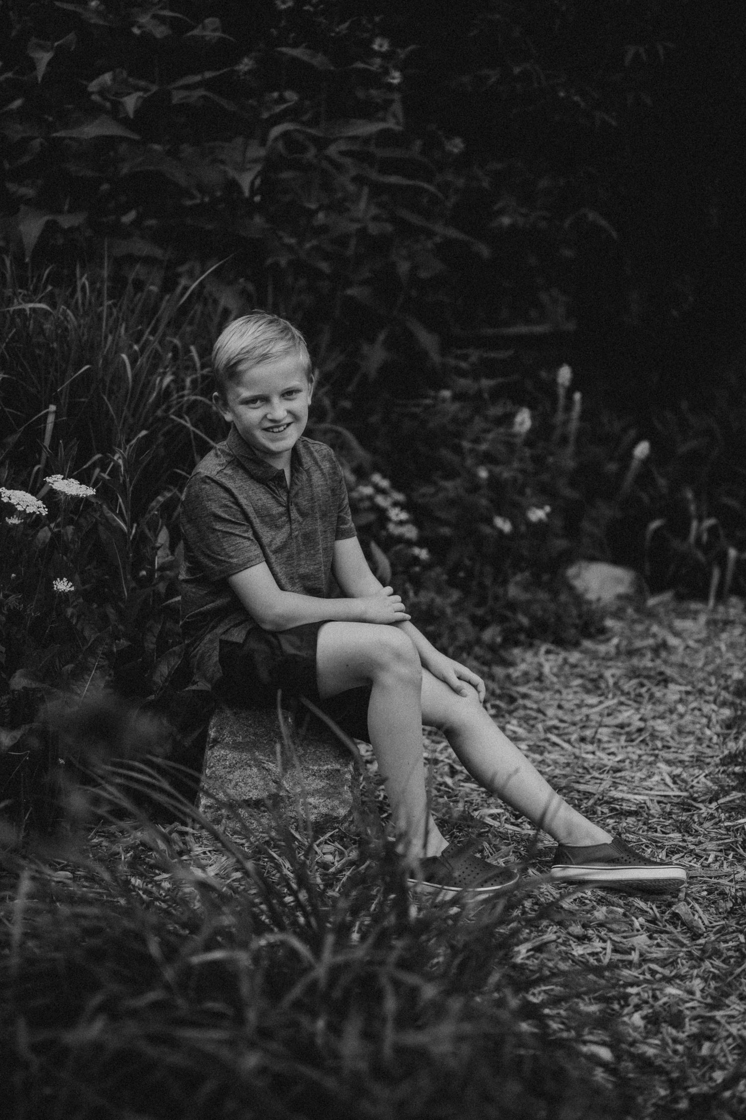 Child sitting on garden path surrounded by flowers and greenery in black and white photo.