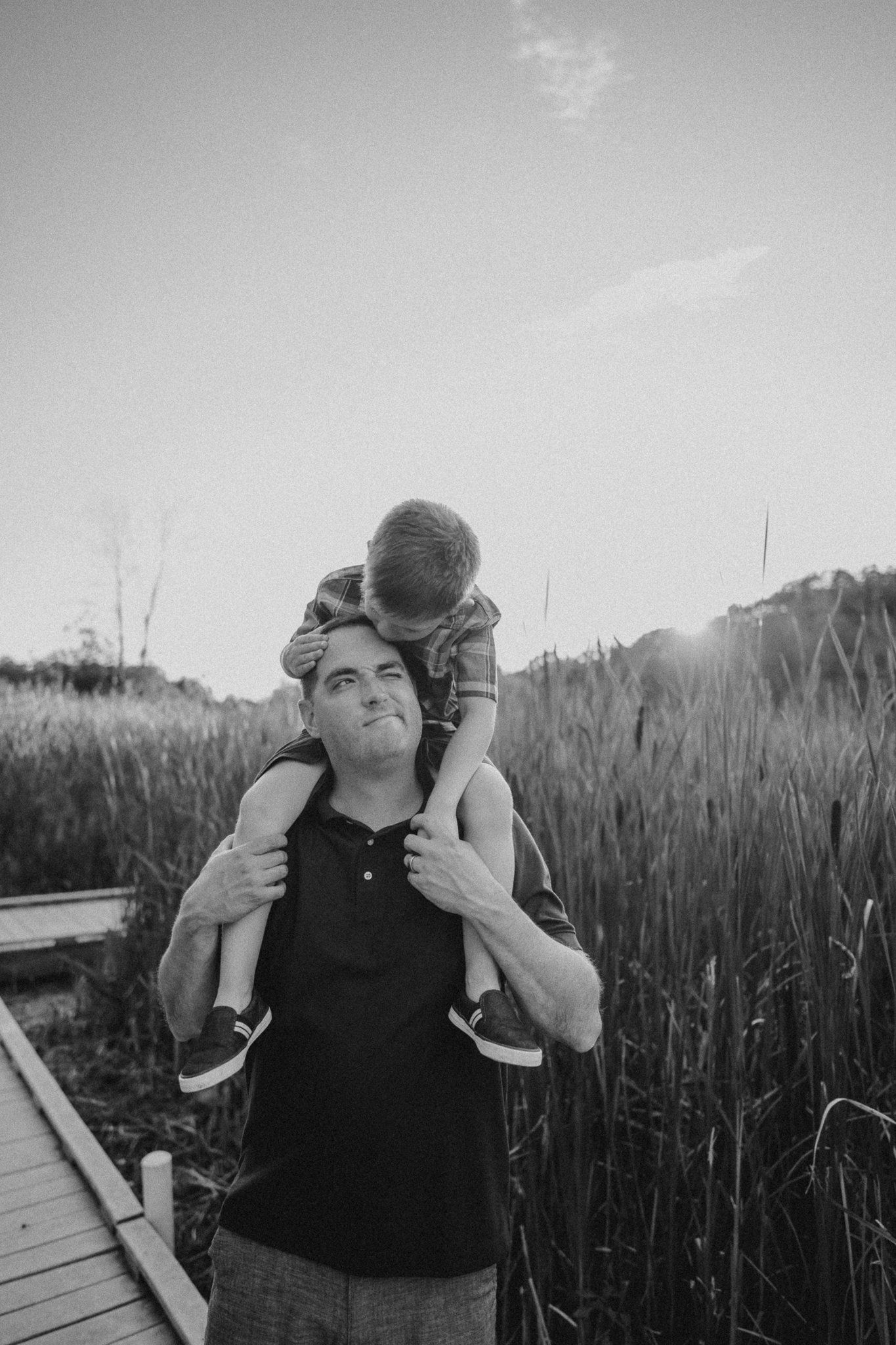 Candid black and white portrait of child riding on parent's shoulders on wooden walkway.