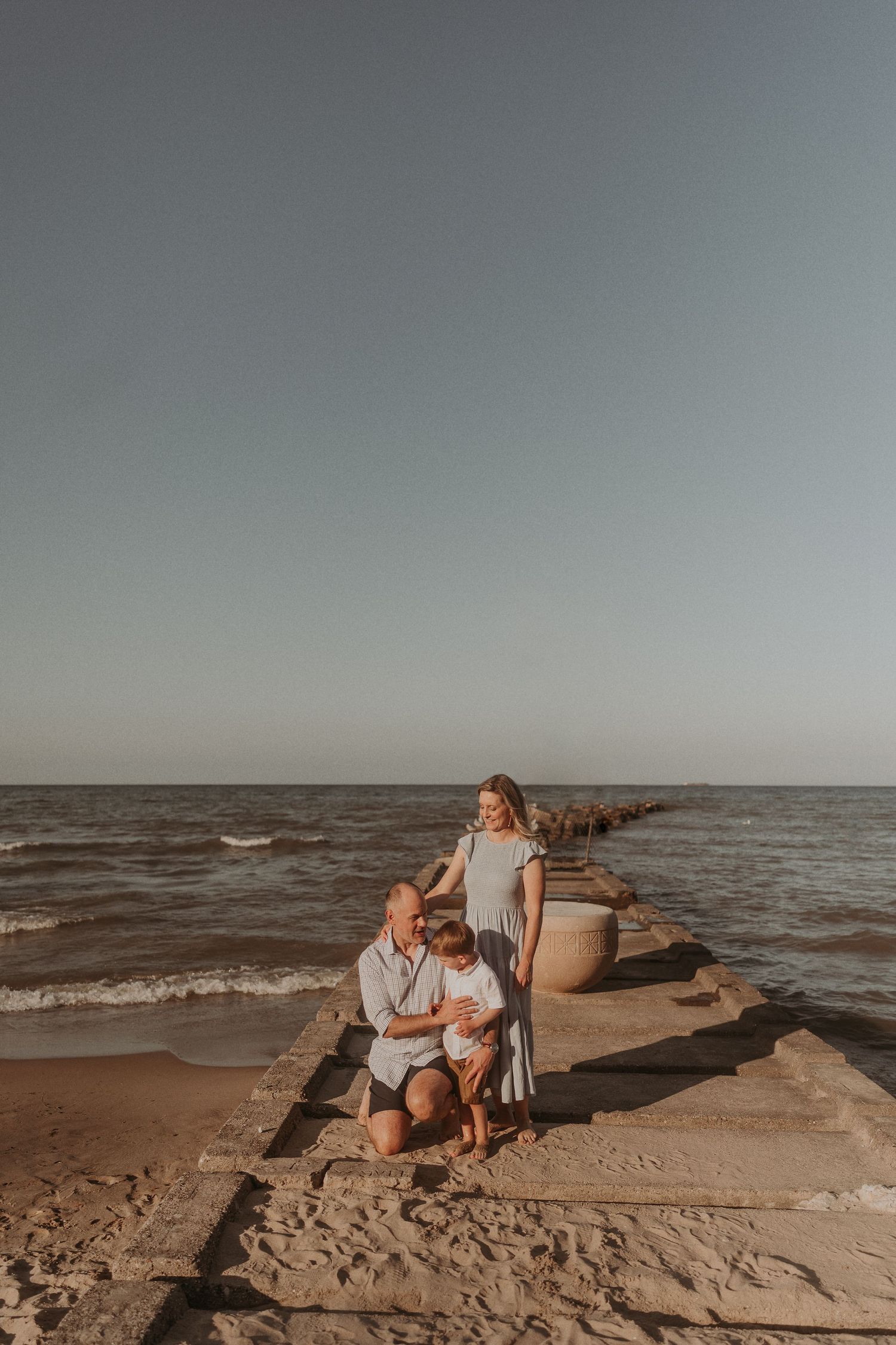 A warm-toned photo of people sitting on a stone pier extending into the ocean at sunset.