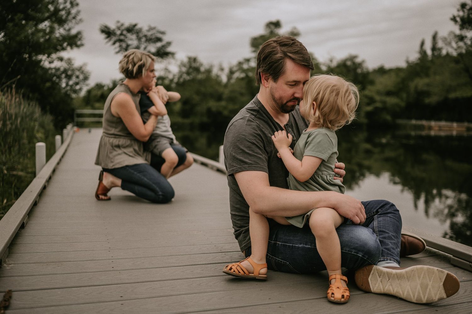 A tender family scene unfolds on a lakeside boardwalk during golden hour.