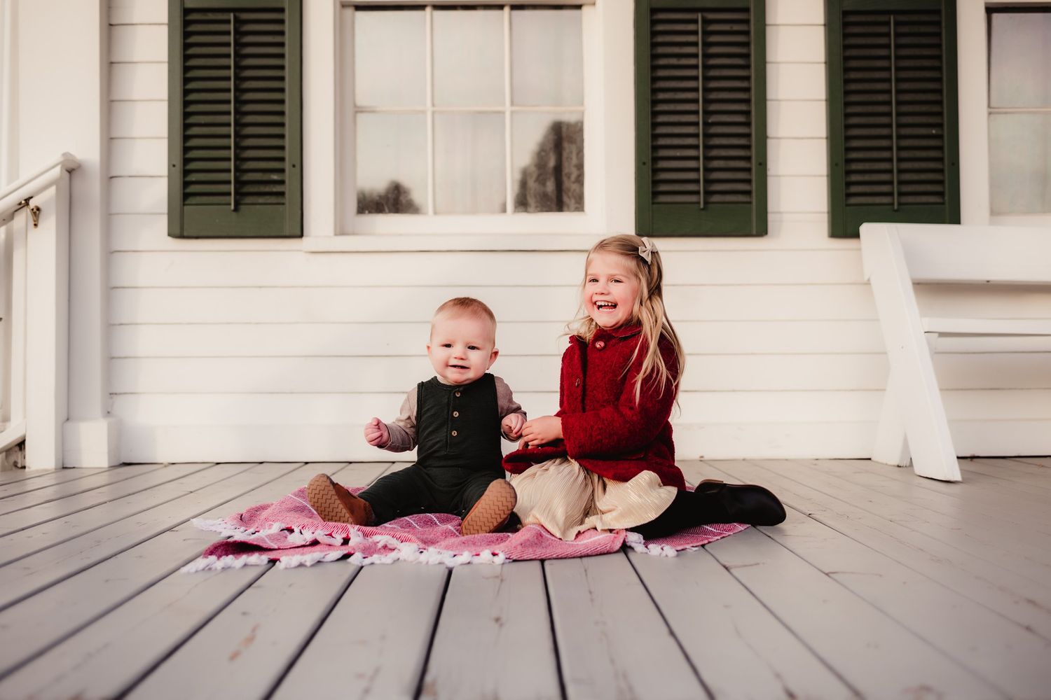 Children sitting together on pink blanket on white porch with green shutters.