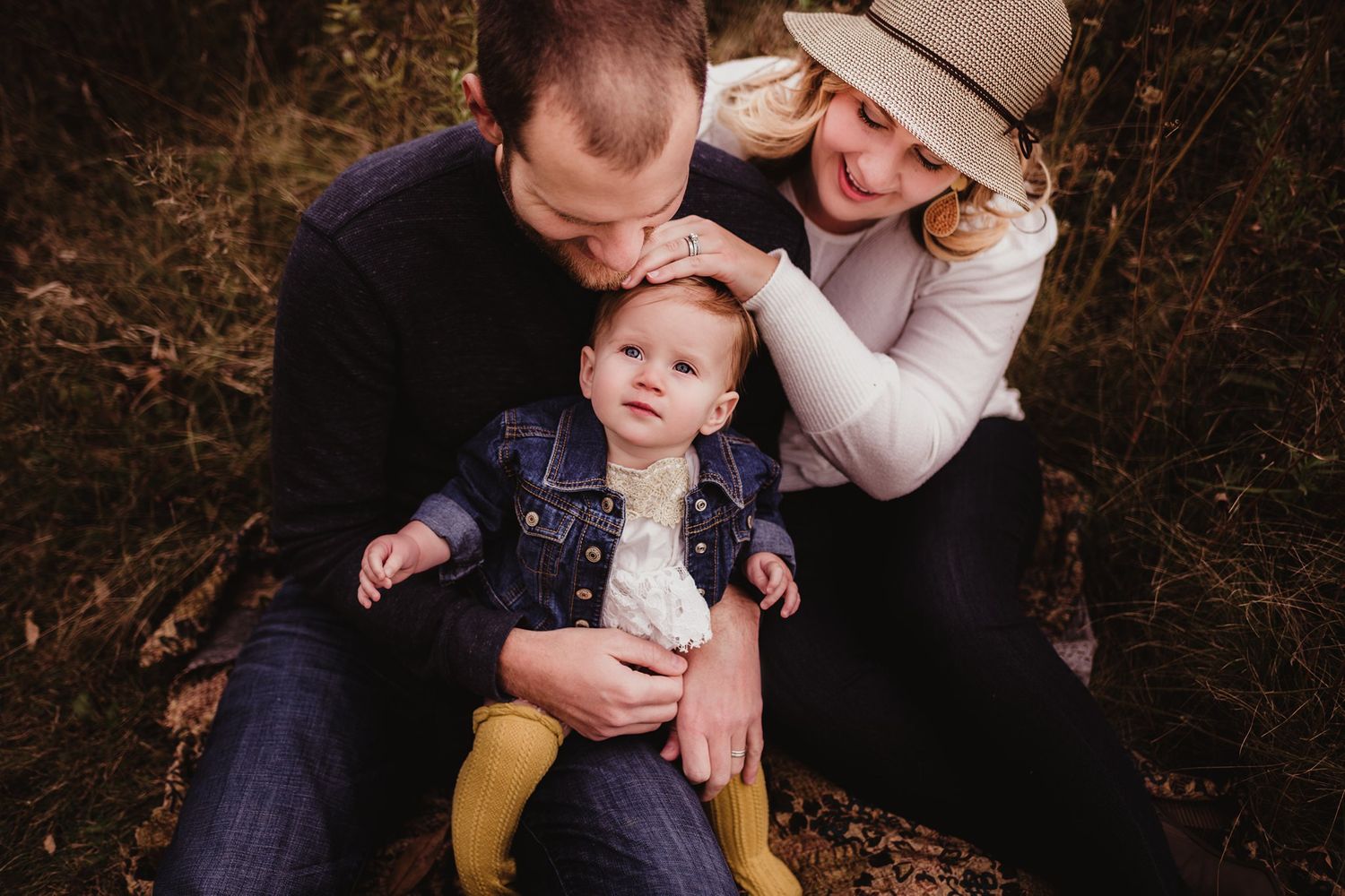 Intimate family portrait outdoors with baby sitting between parents in casual setting.