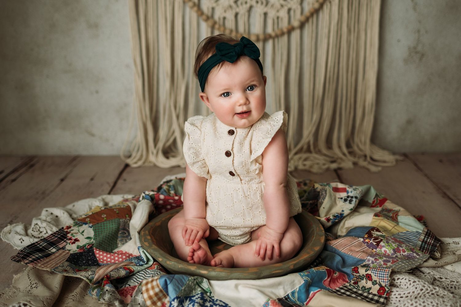 Baby wearing cream colored romper sits in wooden bowl surrounded by vintage quilt pieces.