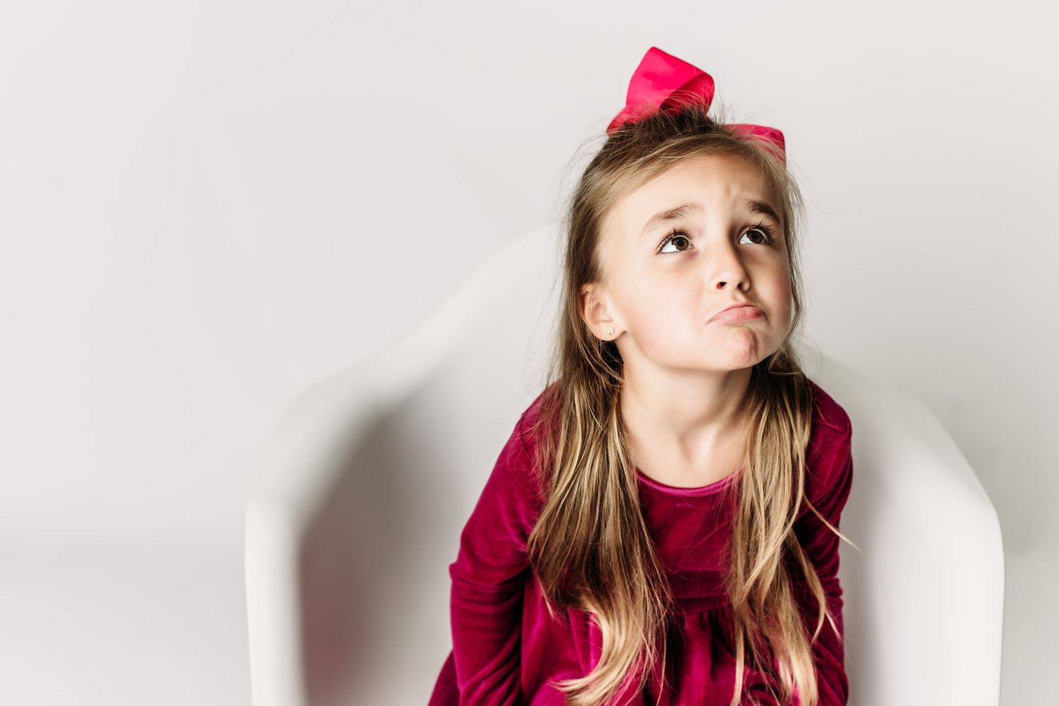 Child in burgundy dress with pink bow making silly face while sitting.