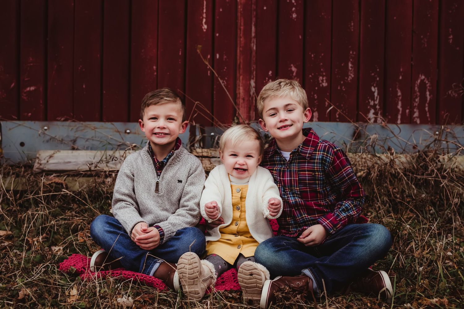 Three young siblings sit together on the ground in front of a red barn for a casual portrait.
