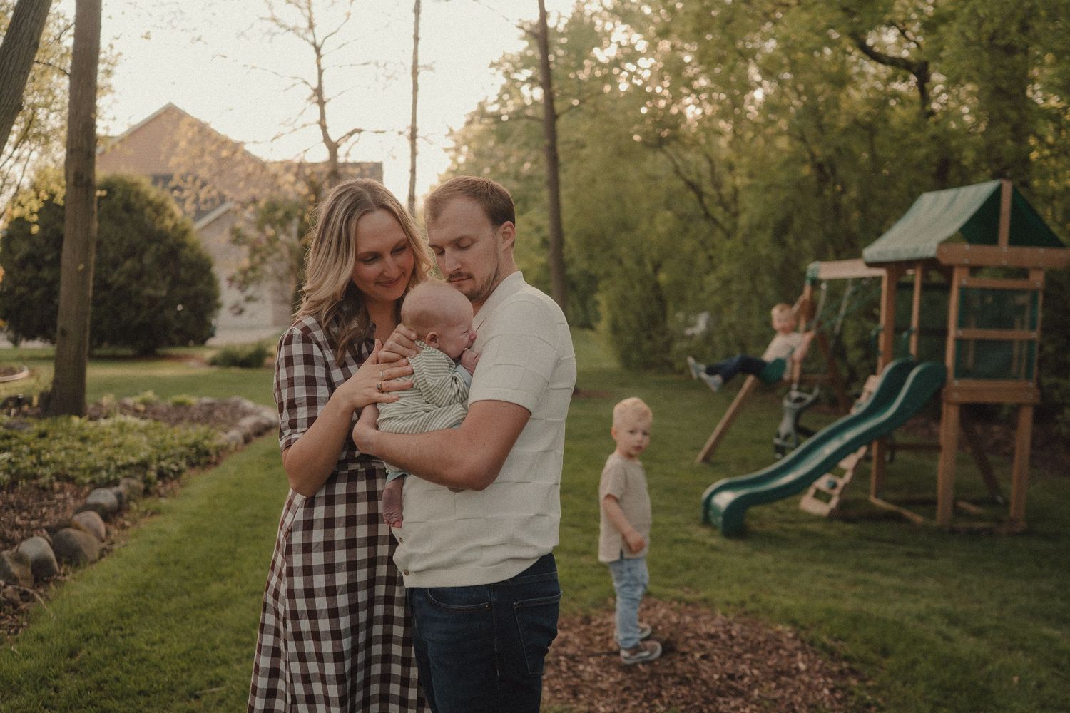 Parents lovingly holding their newborn in a lush backyard while an older child plays near a wooden swing set.