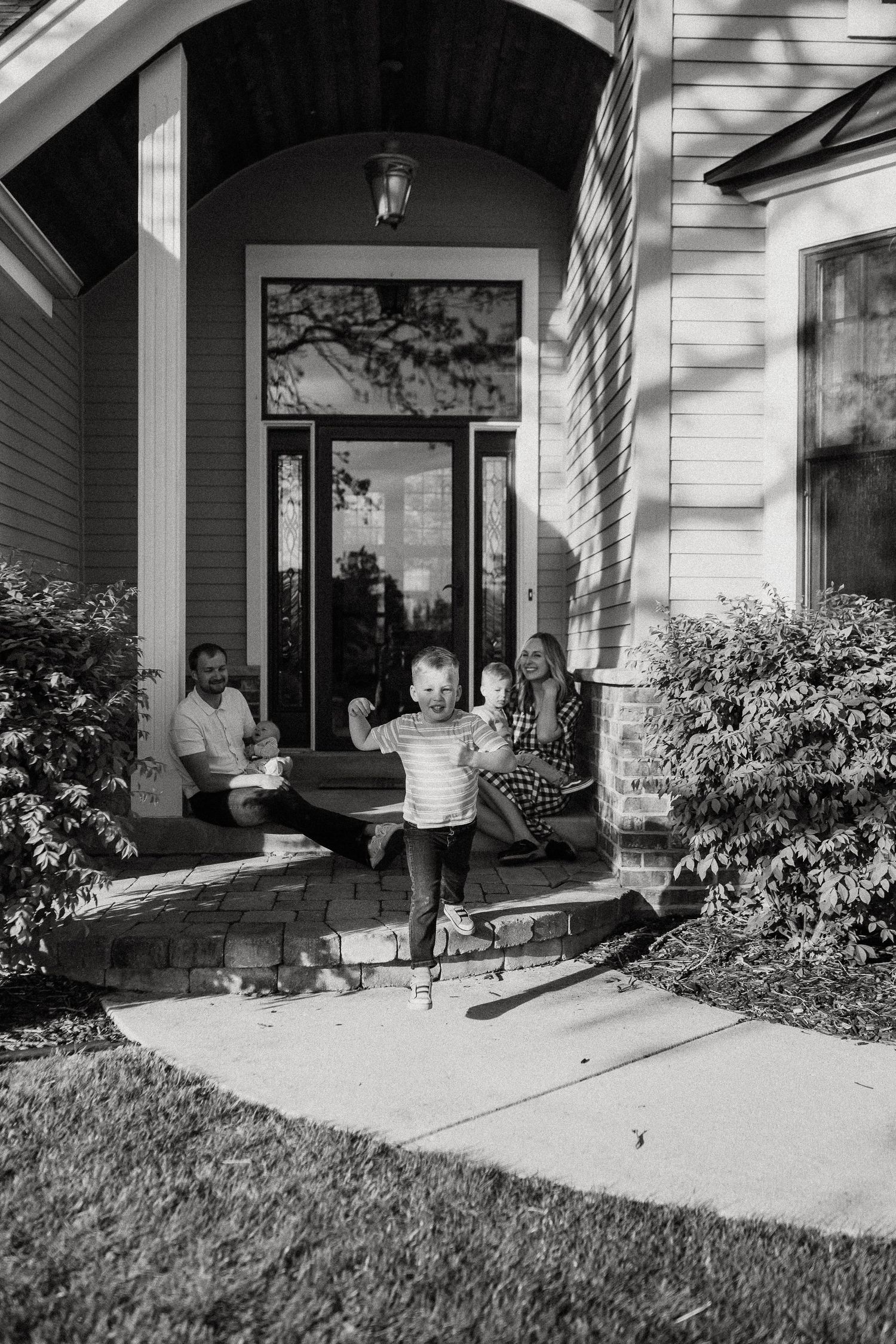 Family relaxing on front porch steps while young child runs down sidewalk in black and white photo.