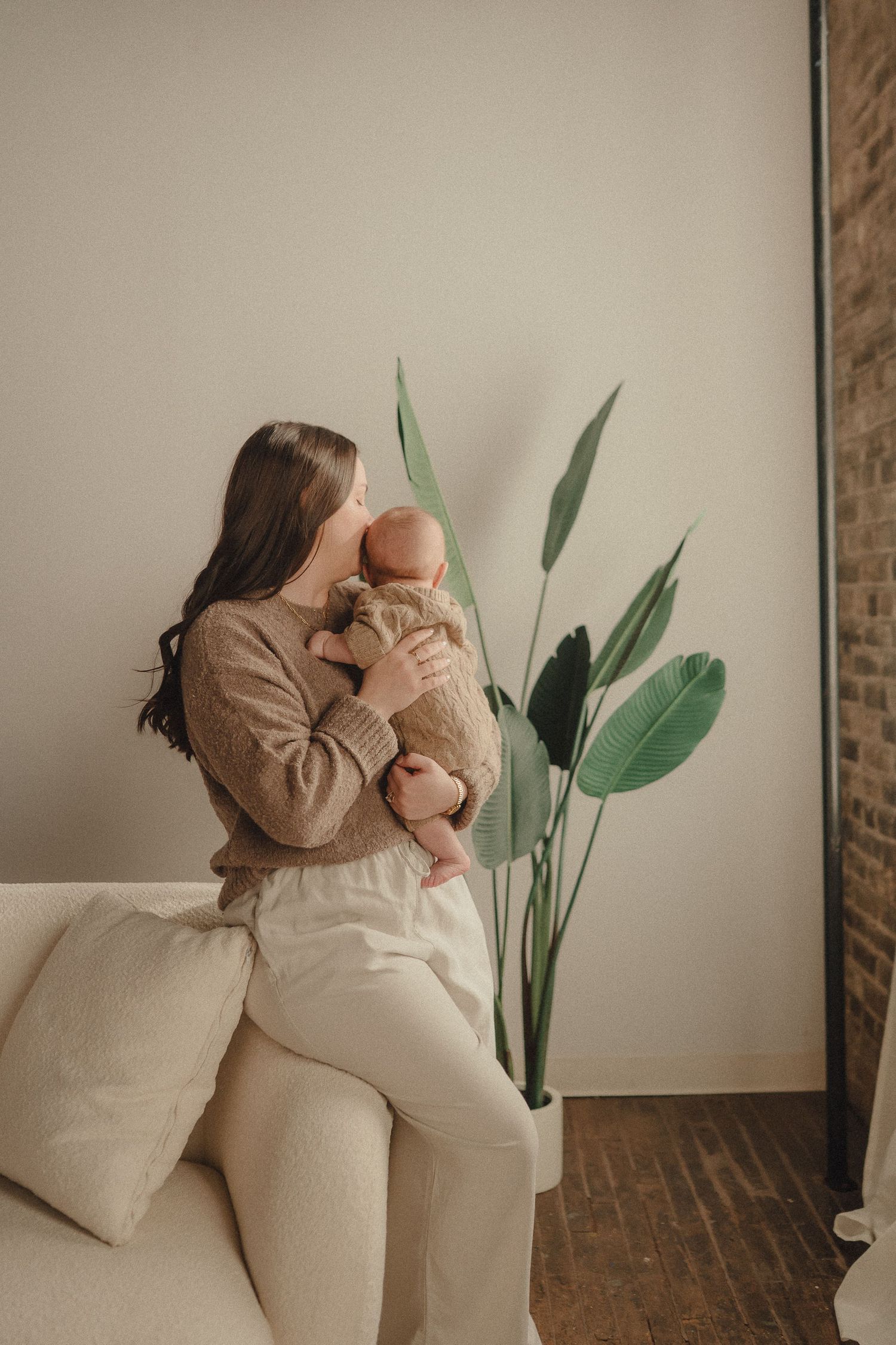 A tender moment between parent and baby in a minimalist room with a large potted plant against a neutral wall.