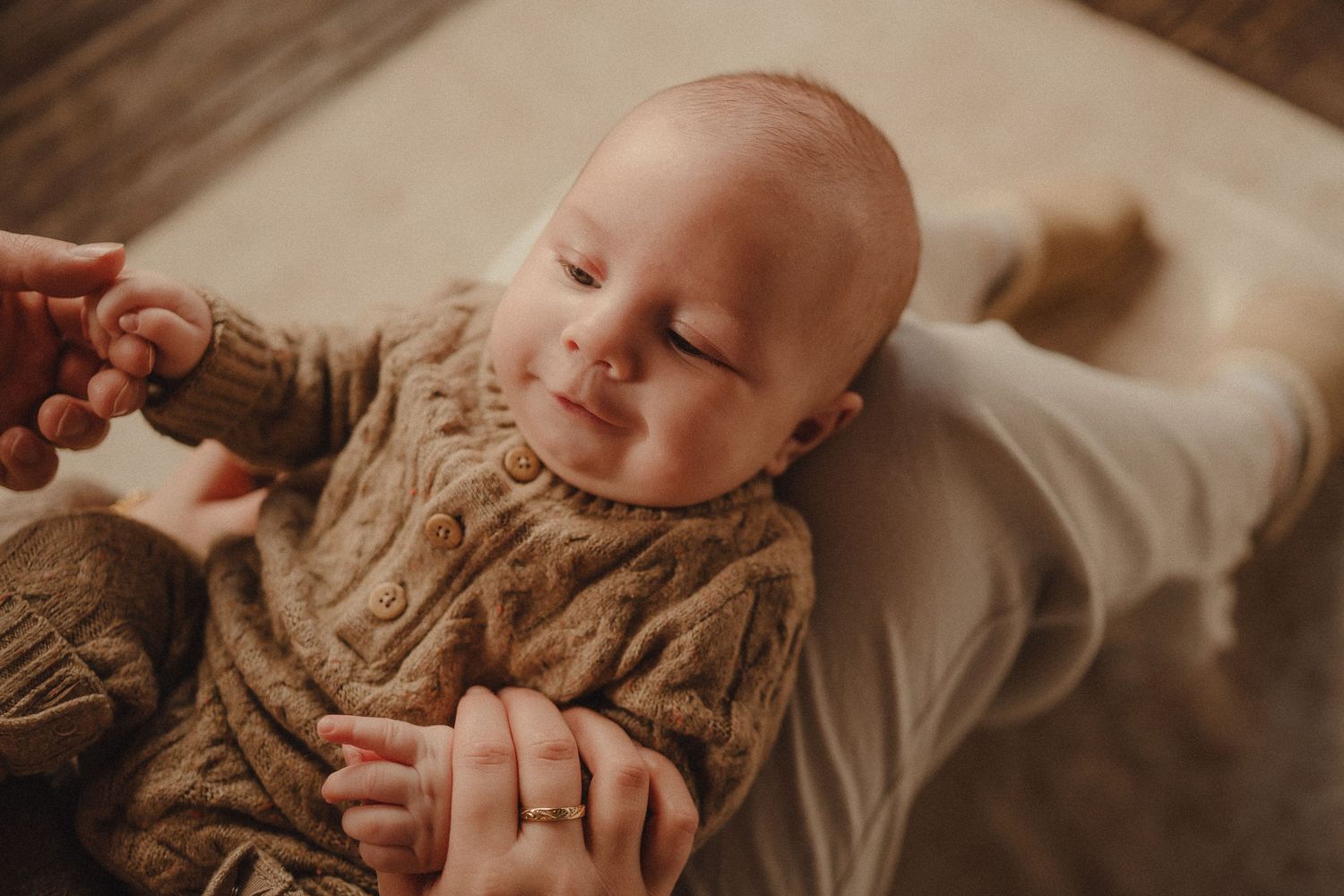 A baby in a brown knit sweater smiles while lying on a white surface.