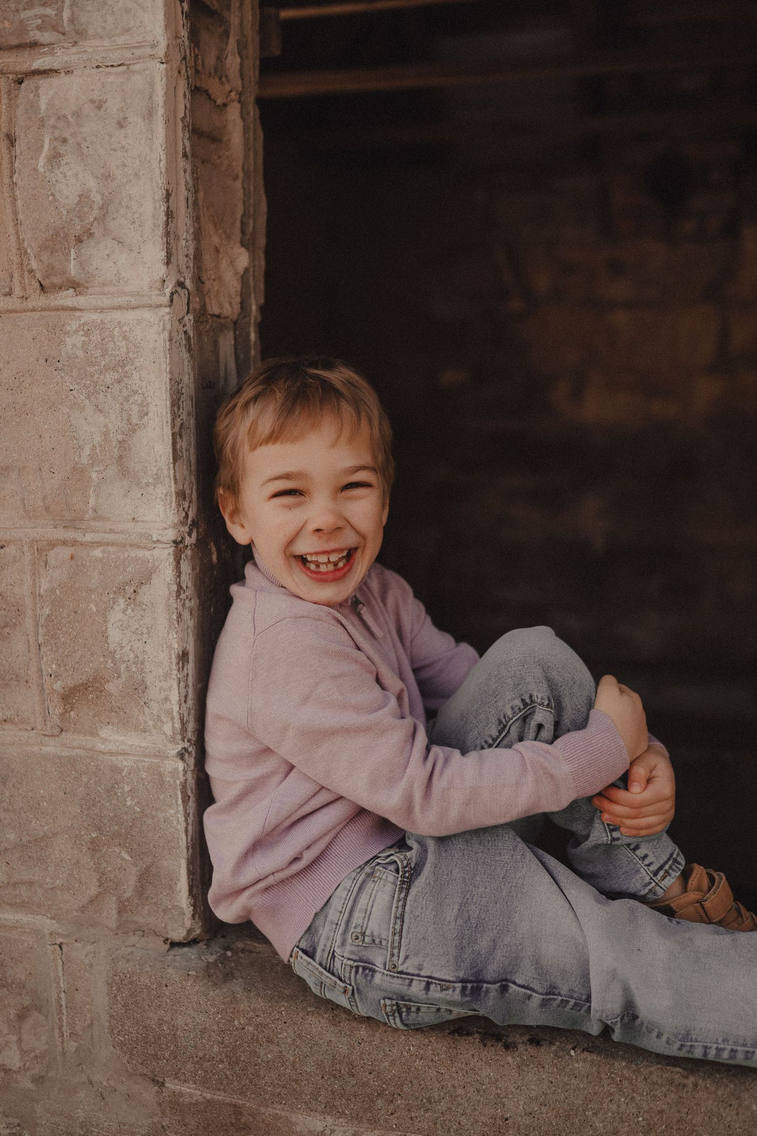 A child sits in a stone doorway wearing a pink sweater and jeans while smiling brightly.