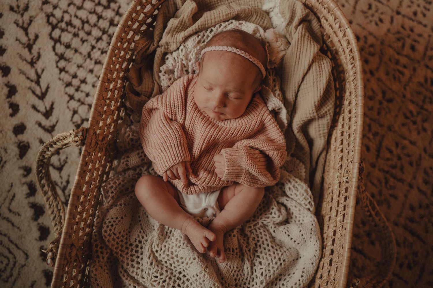Newborn baby sleeping peacefully in a cozy woven basket with textured blankets and warm lighting.