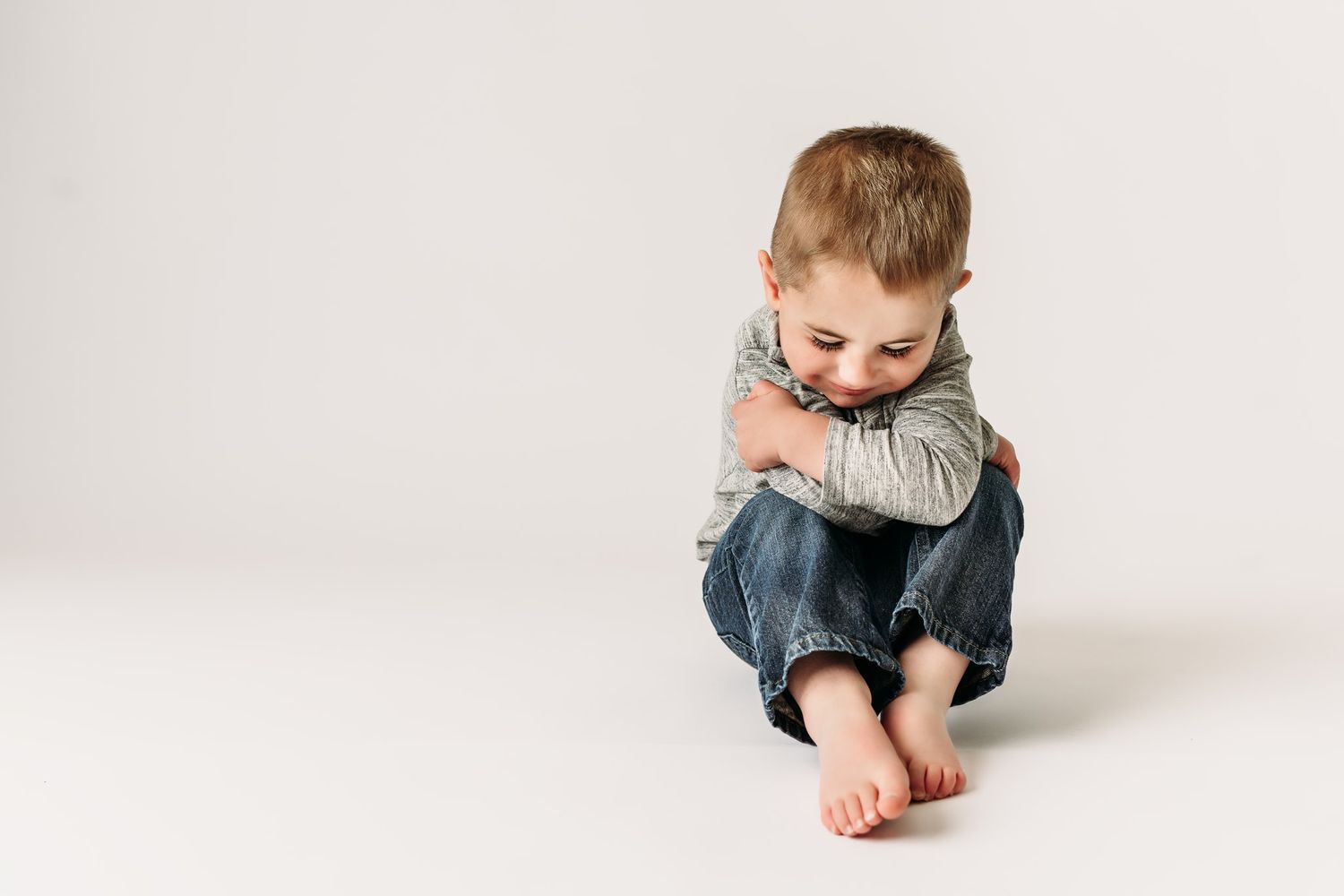 Child in casual wear hugs knees while sitting on white backdrop.