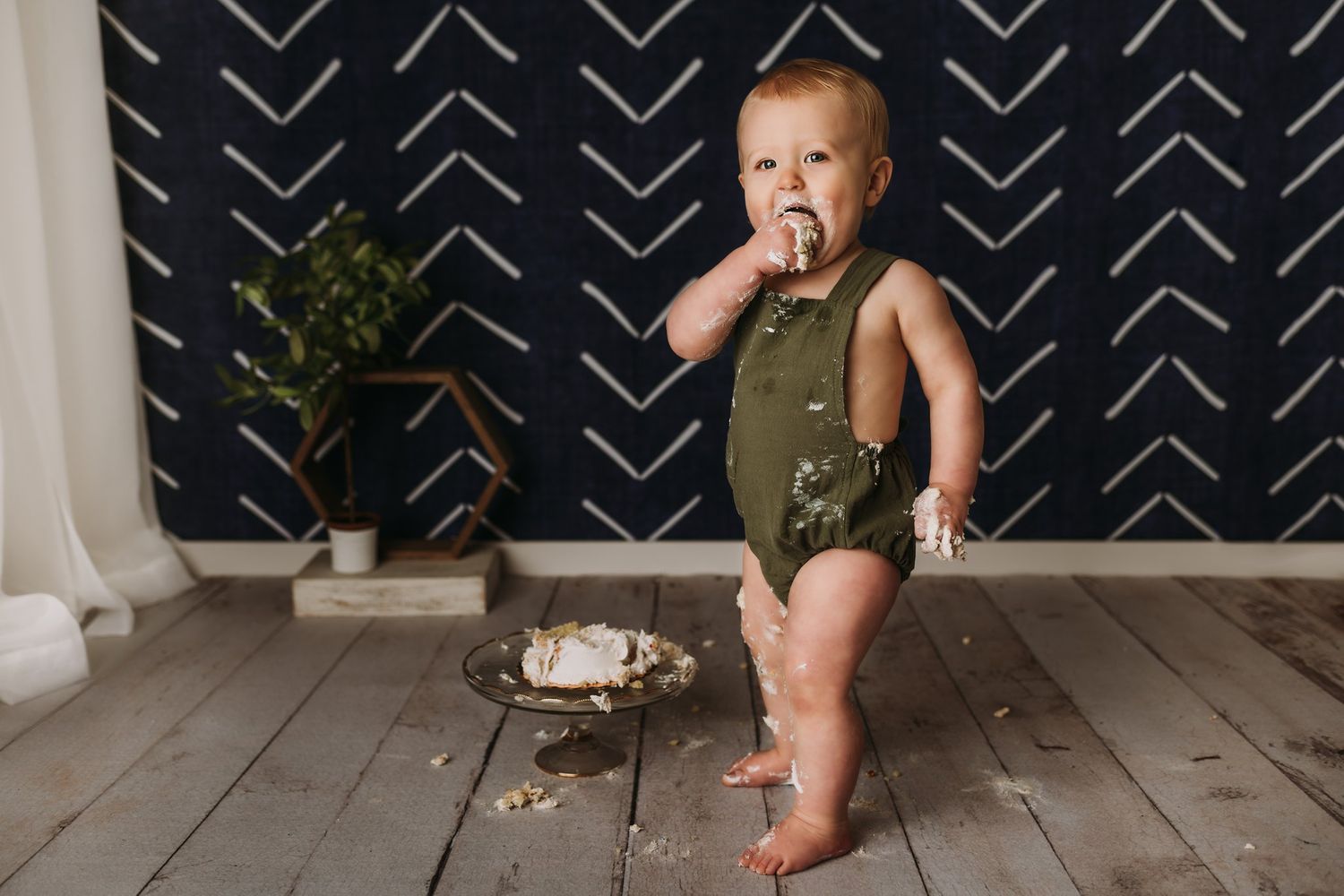Baby in green outfit standing near partially eaten cake with geometric wall background.
