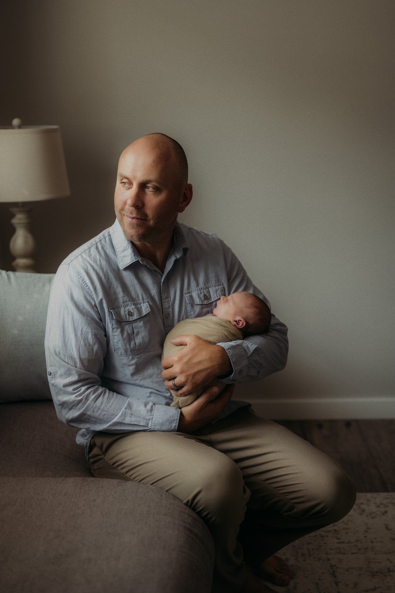 Indoor lifestyle portrait of an adult holding a sleeping newborn in a grey room with soft lighting.
