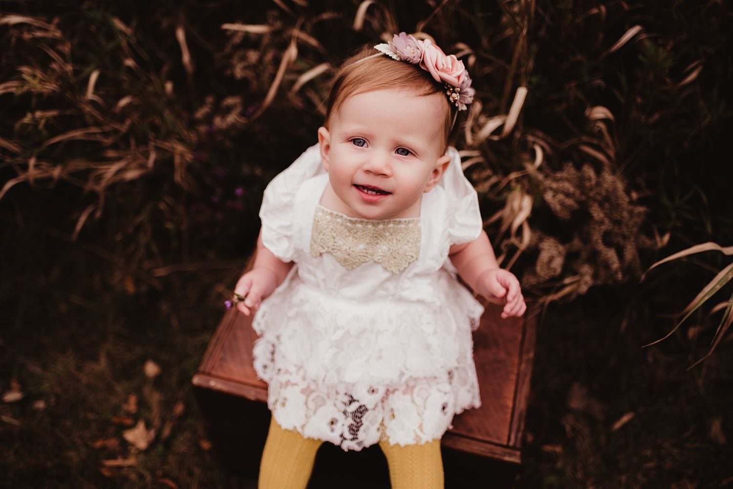 A small child in a white lace dress and yellow tights sits on a wooden chair outdoors.