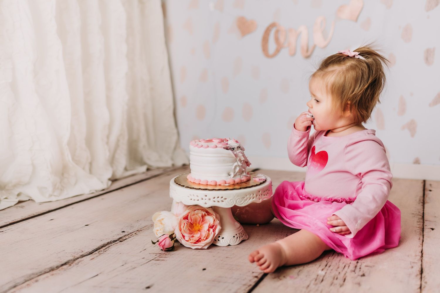 Pink and white themed celebration setup with cake and floral decorations on wooden floor.