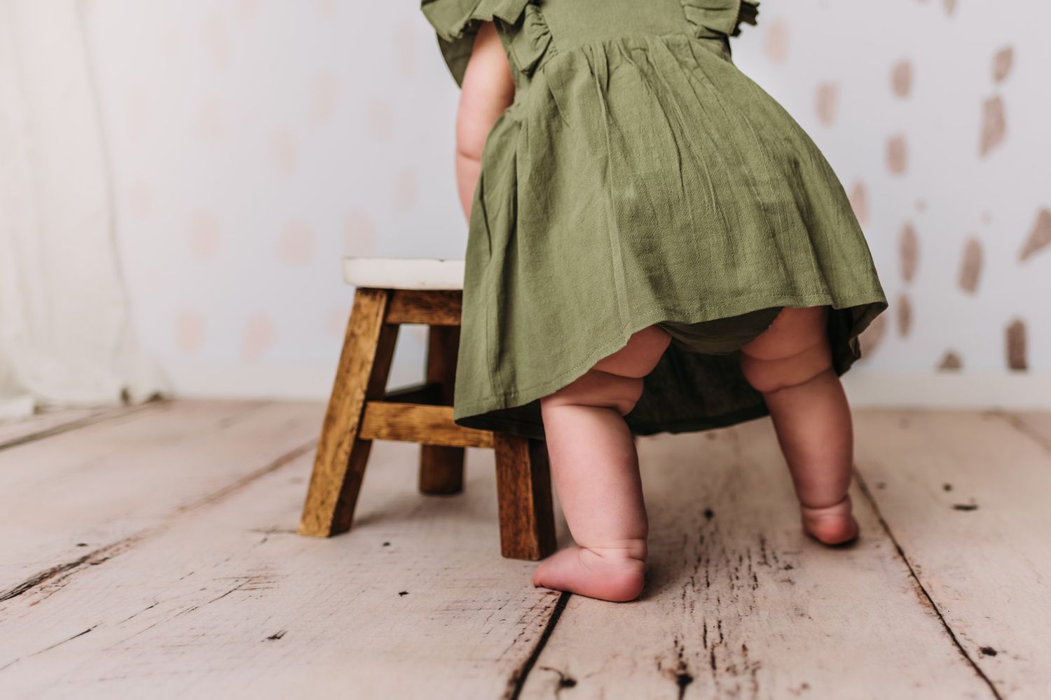 Close-up view of olive green dress and wooden stool details on white wooden floor.