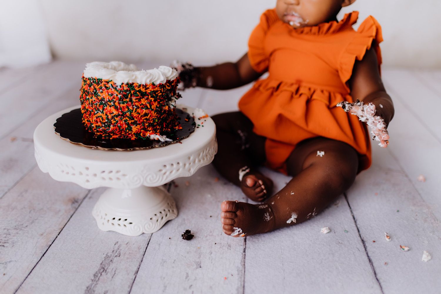 Decorated chocolate cake on white pedestal next to baby in orange dress during cake smash session.