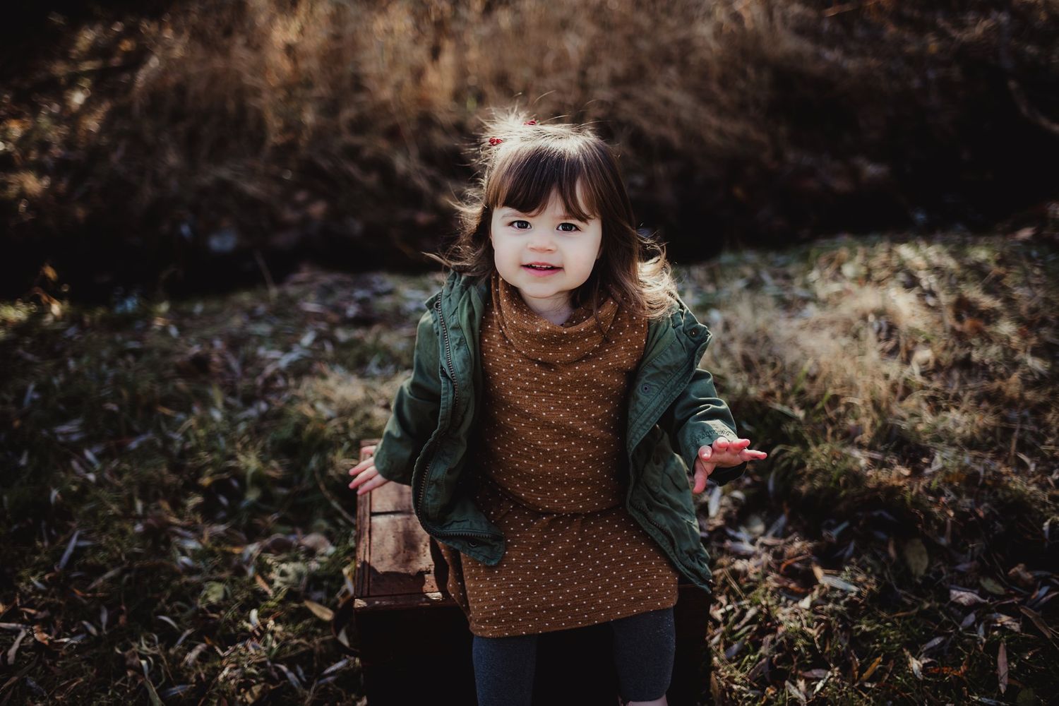A small child in winter clothing poses outdoors against an autumn background.