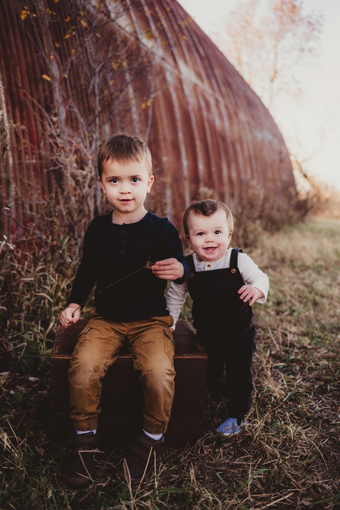 Two children sitting together on a wooden bench against a rustic red barn wall in casual attire.