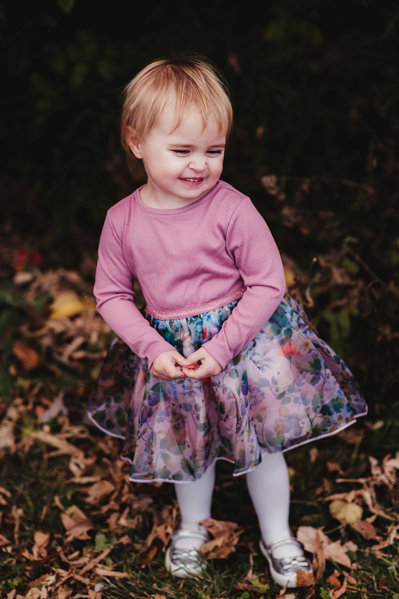 Toddler wearing pink sweater and floral skirt smiles while playing in autumn leaves.