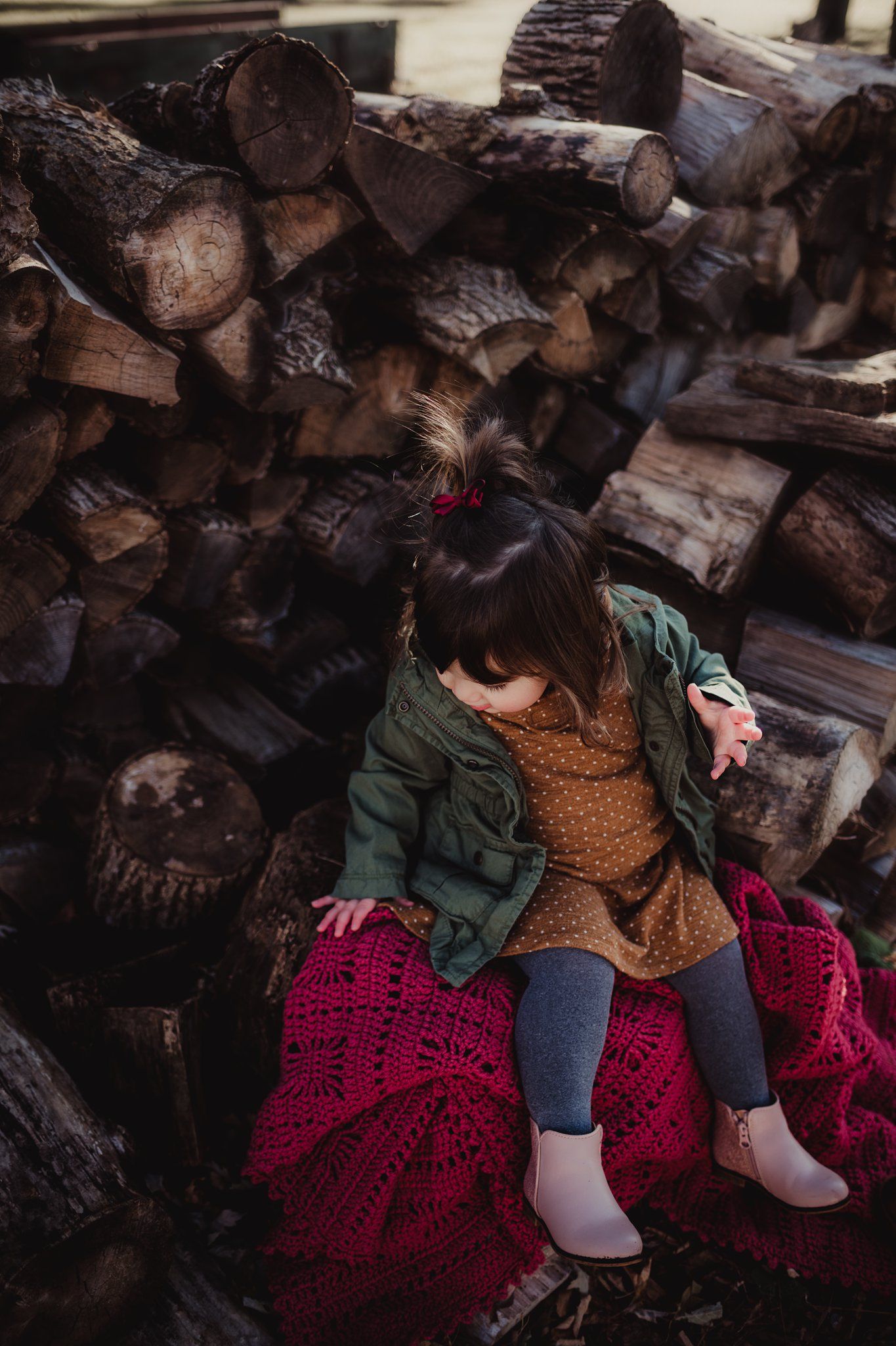 A small child in outdoor wear sits on a red blanket against stacked firewood.