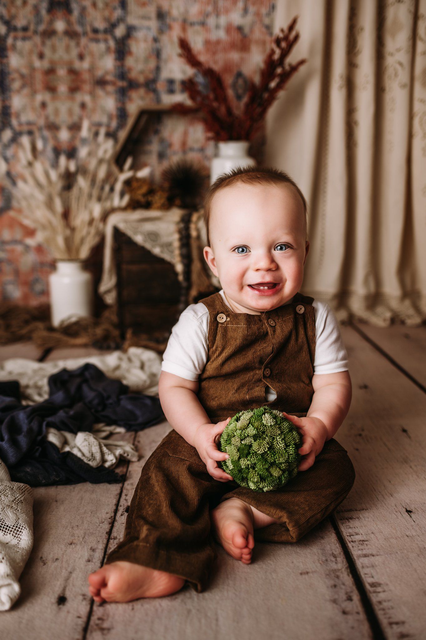 Smiling with green prop in brown overalls against vintage brick and fabric backdrop.
