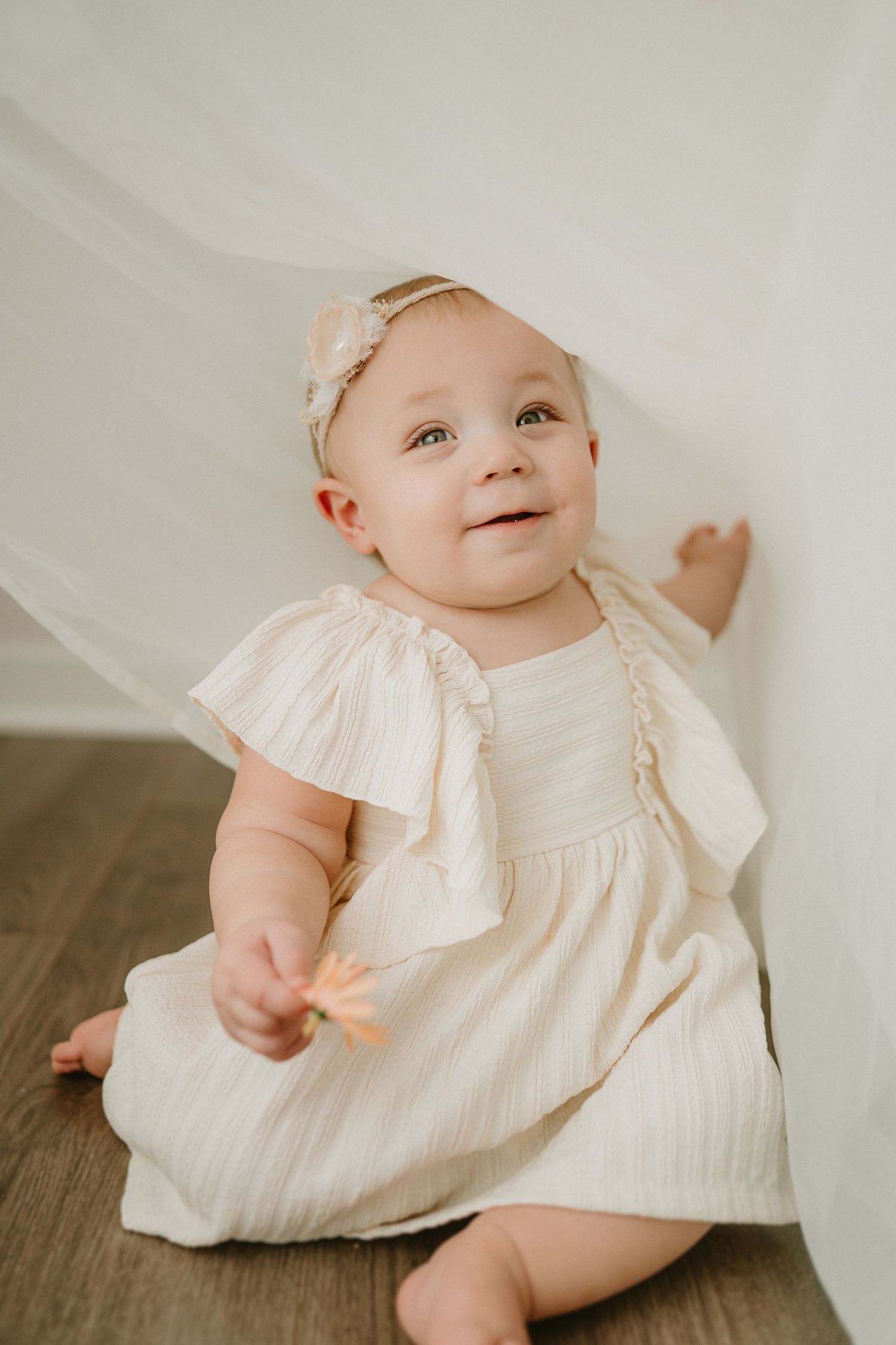 Baby wearing a white cotton dress and matching headband sits on hardwood floor with a happy expression.