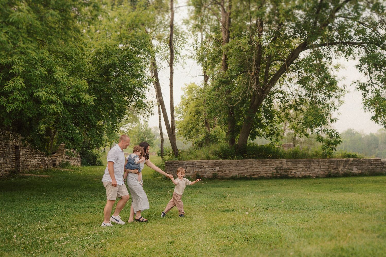 A joyful outdoor scene shows figures dancing together on a green lawn with tall trees and stone wall in background.