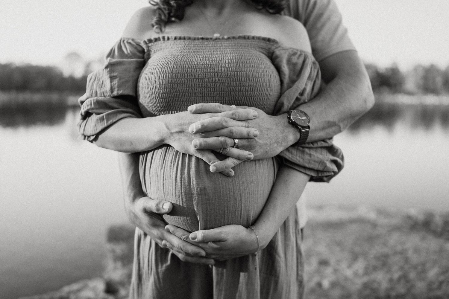 Black and white close-up of hands tenderly holding pregnant belly by waterfront.