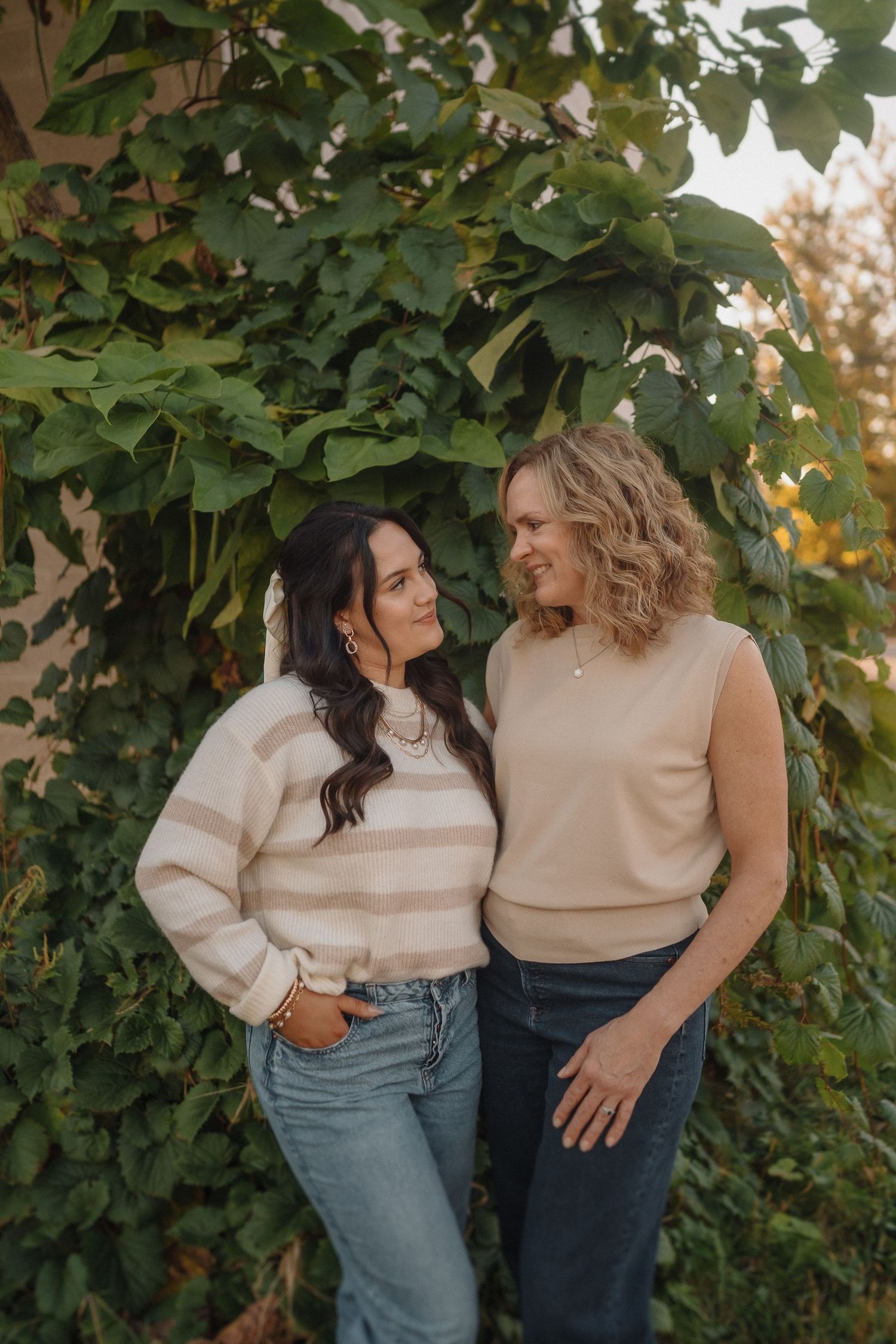 Two friends in cozy sweaters and jeans pose together against lush green foliage in an outdoor setting.