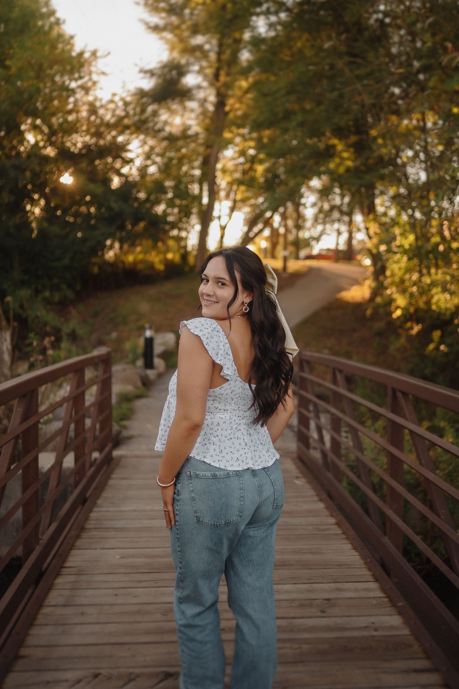 A person in white top and jeans poses on a wooden bridge at sunset in a wooded area.