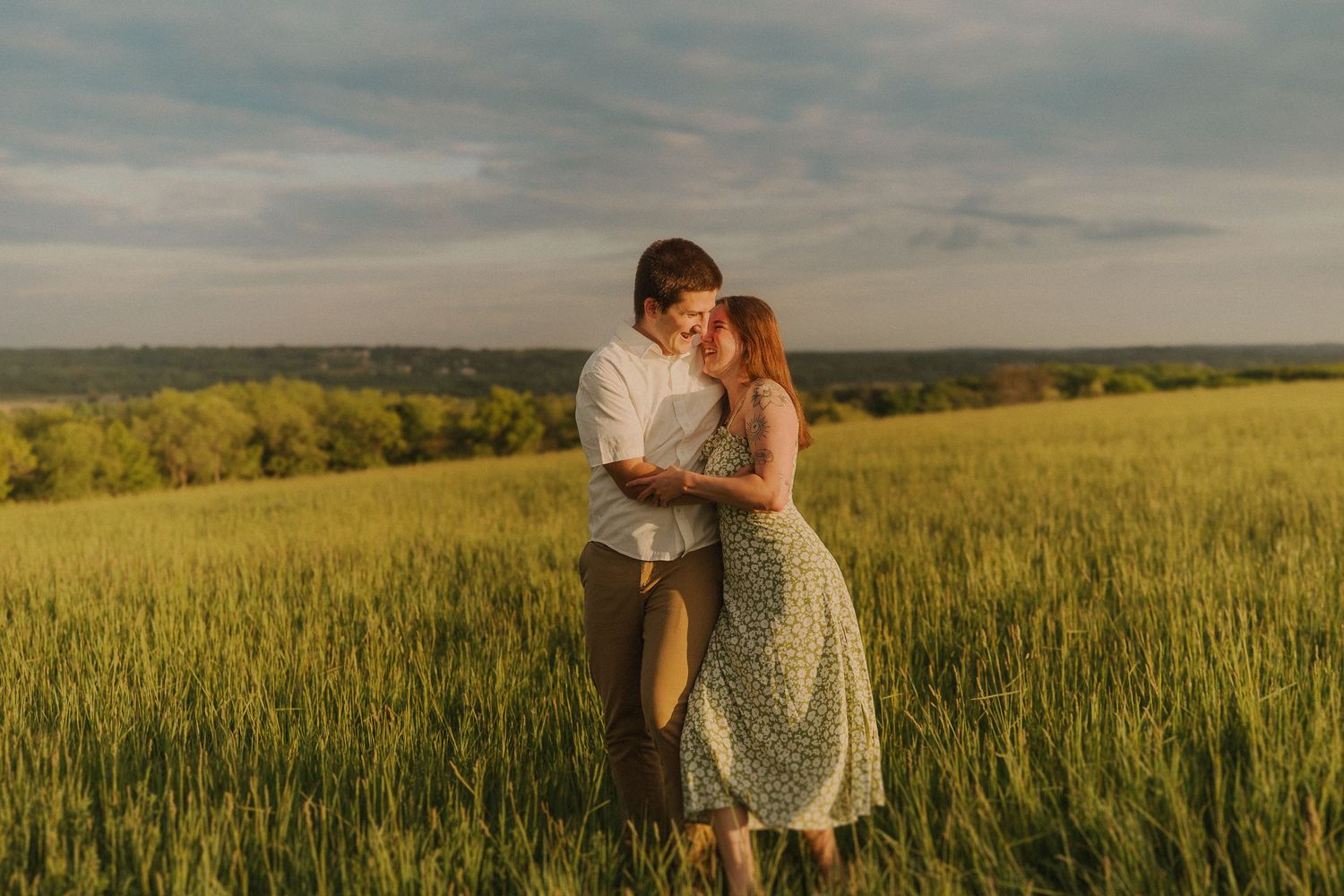 A romantic couple embraces in a golden wheat field at sunset with rolling hills in the background.