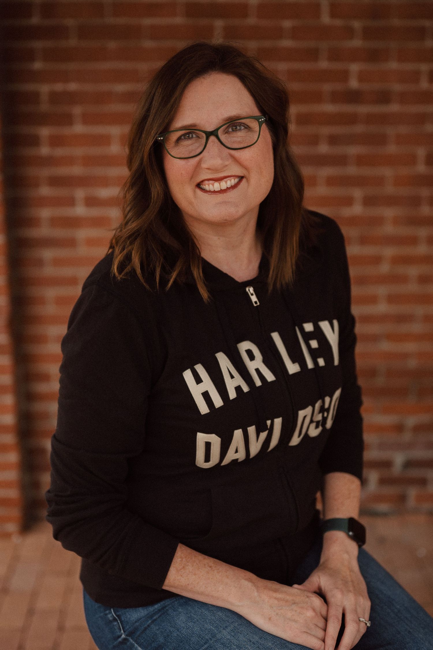 Six professional headshots of someone wearing a black sweatshirt and glasses against a brick wall background.