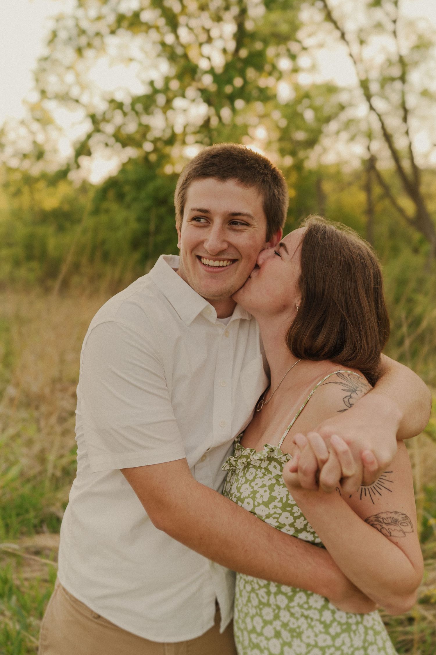 A couple shares an intimate moment laughing and embracing in a sunlit field with trees in the background.