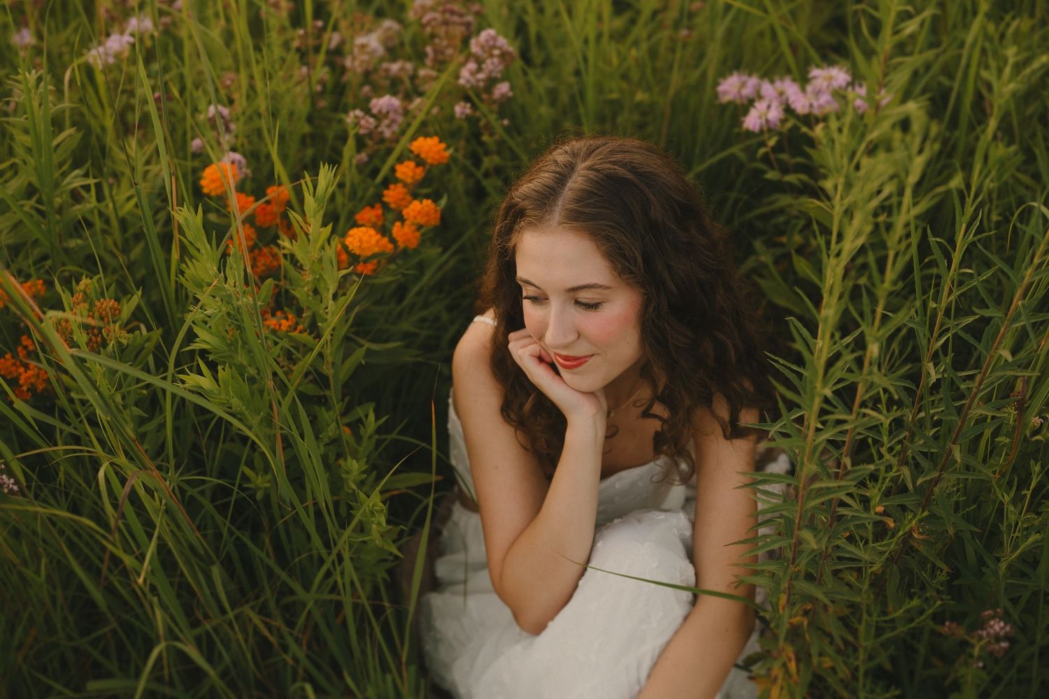 A dreamy summer portrait shows someone resting peacefully among orange and purple wildflowers in tall grass.