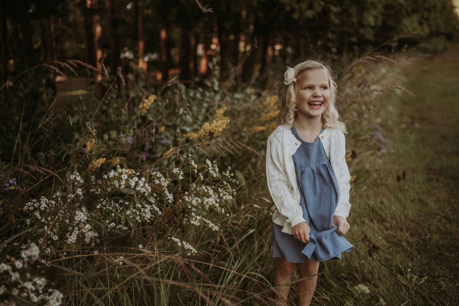 Child wearing a blue dress and white sweater smiling brightly in an outdoor setting with natural foliage.